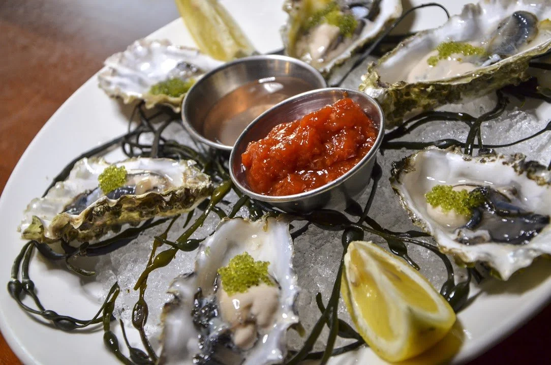 Oysters on the half shell with lemon wedges, mignonette sauce, and cocktail sauce, served on ice with seaweed decorations.