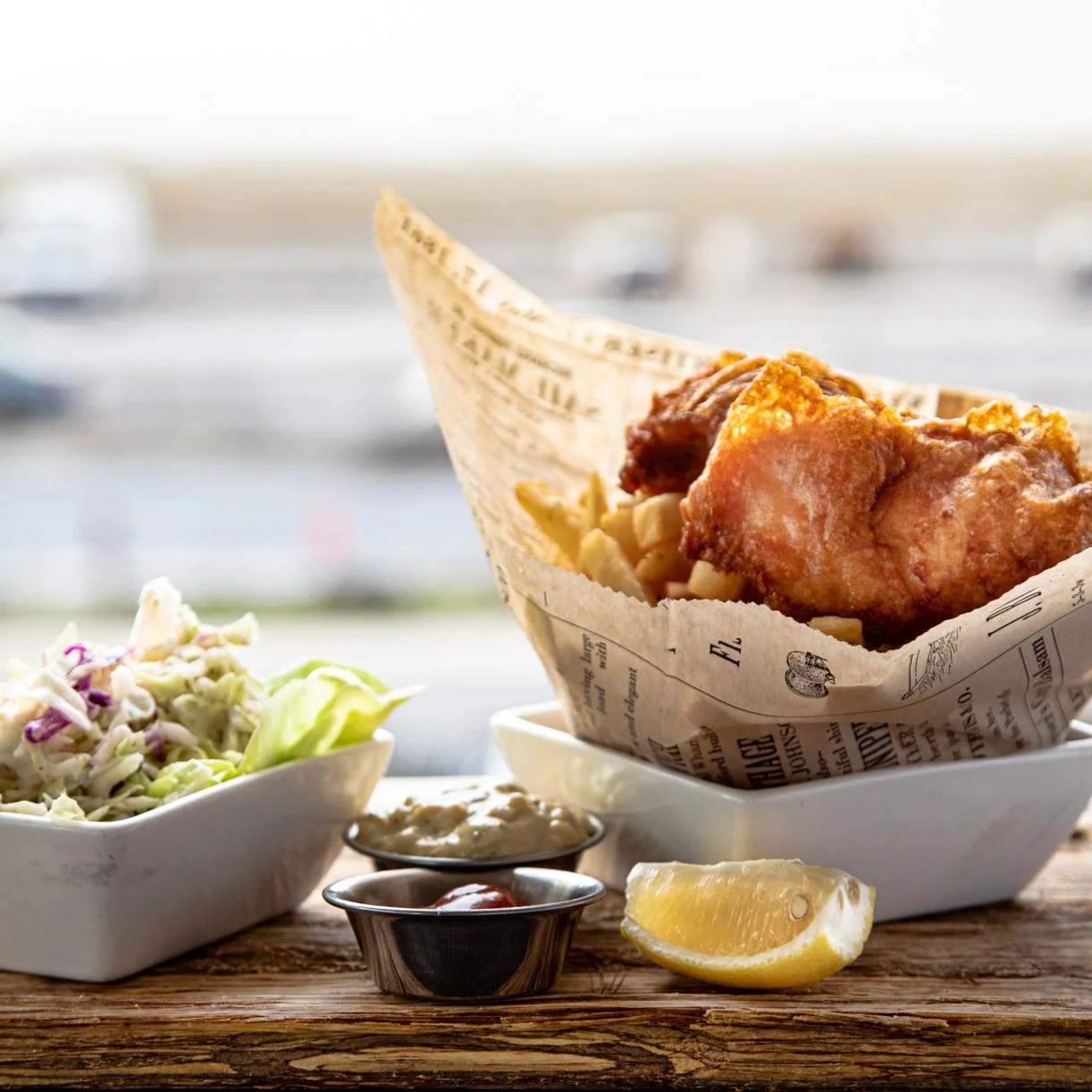 Fried chicken served in a paper-lined basket, with a side of coleslaw, lemon wedge, and small dishes of dipping sauces on a rustic wooden table with a blurred outdoor background.