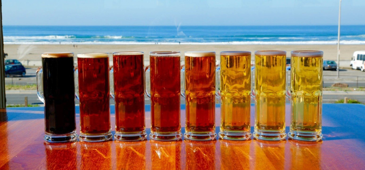 Seven glasses of different types of beer lined up on a wooden table with a beach and ocean in the background.