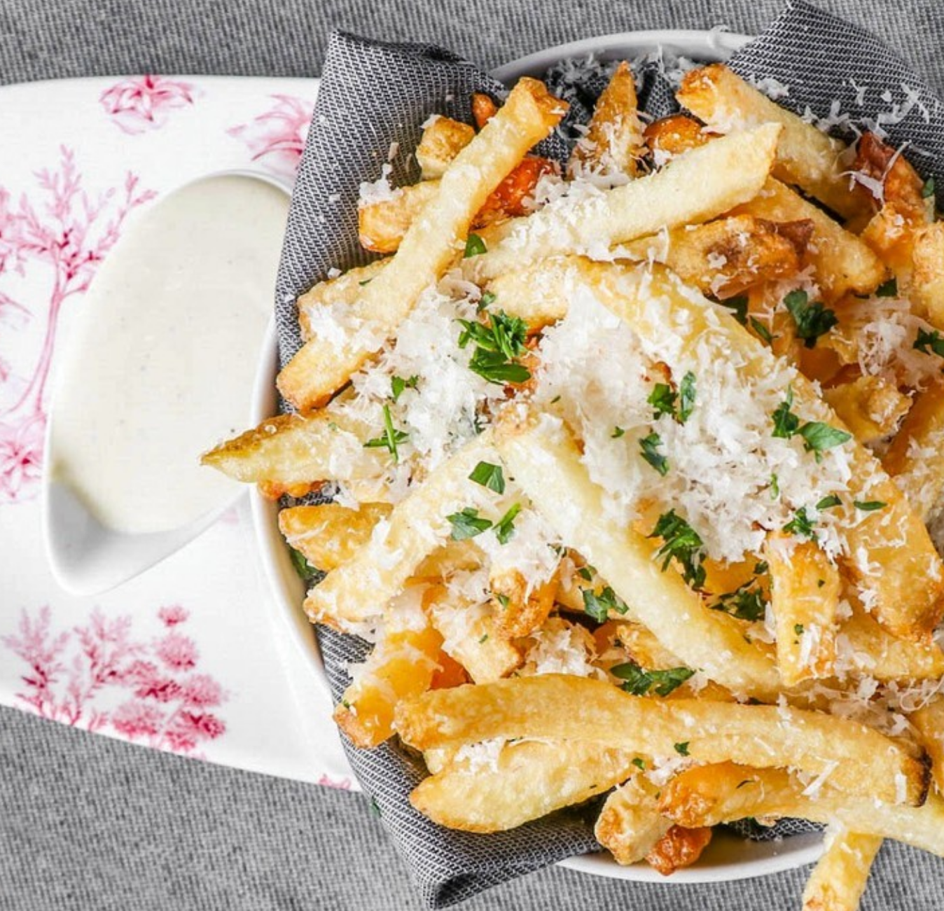 Basket of crispy French fries topped with grated cheese and parsley, with a side of creamy dipping sauce.
