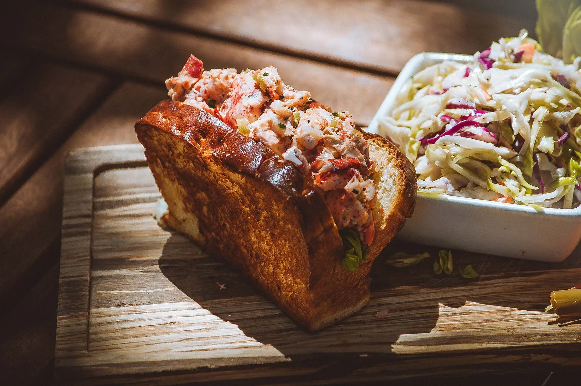 Lobster roll on toasted bun with side of coleslaw in a white square bowl.