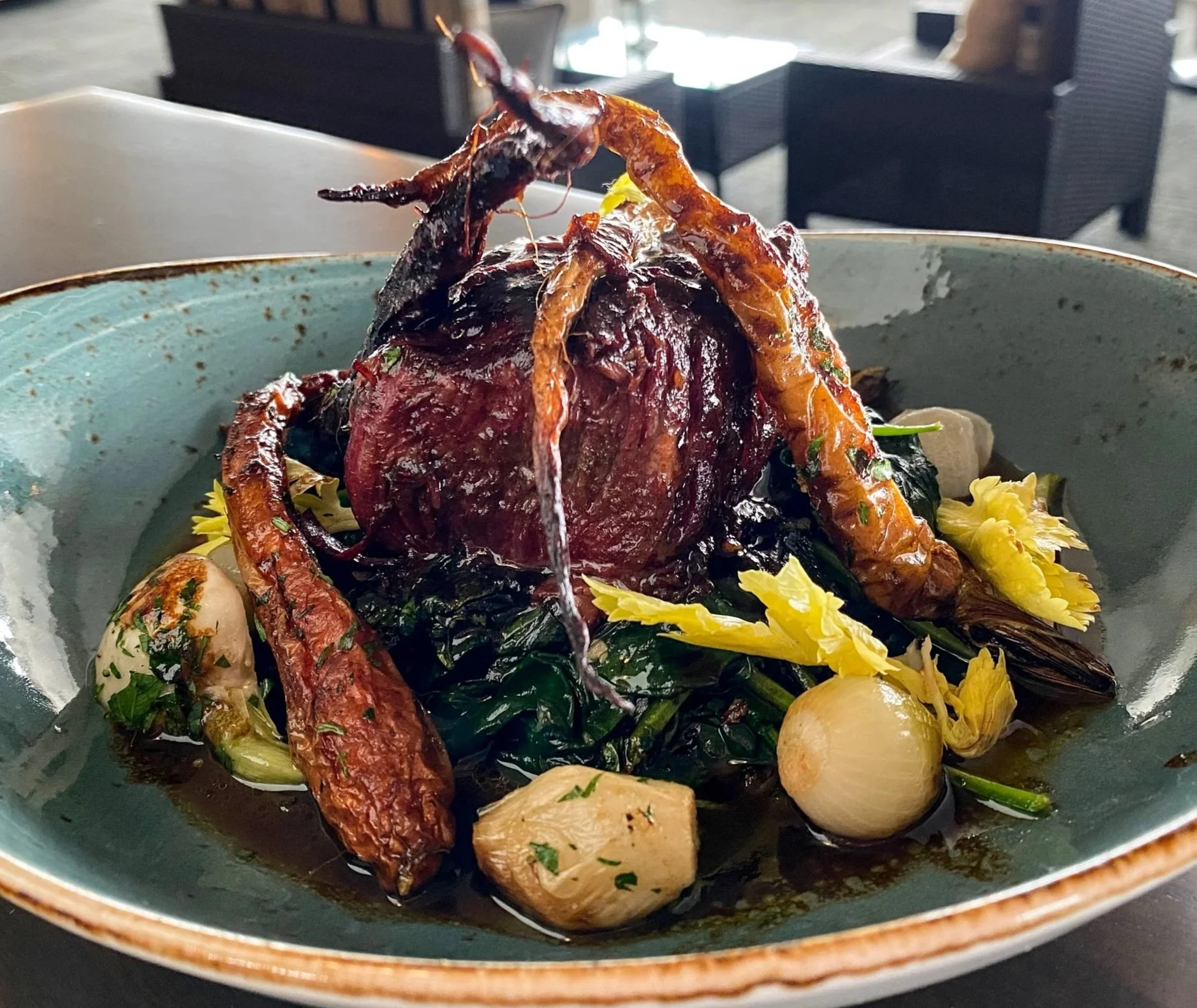 A plated gourmet meal featuring a cooked lobster with a dark shell, accompanied by boiled potatoes, leafy greens, and yellow flowers, in a blue ceramic bowl.