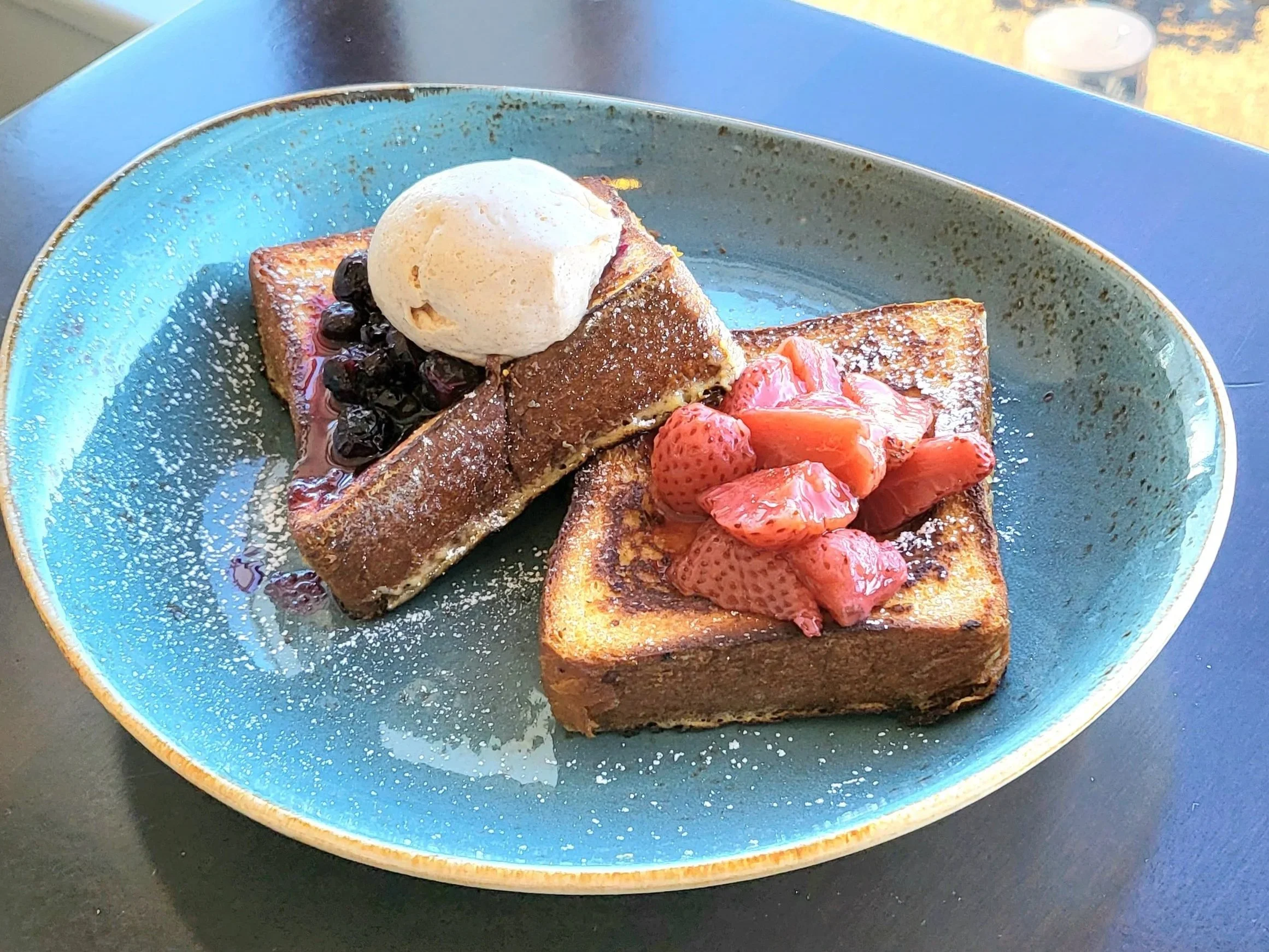 Slice of French toast topped with strawberries, blueberries, a scoop of vanilla ice cream, and powdered sugar on a blue ceramic plate.