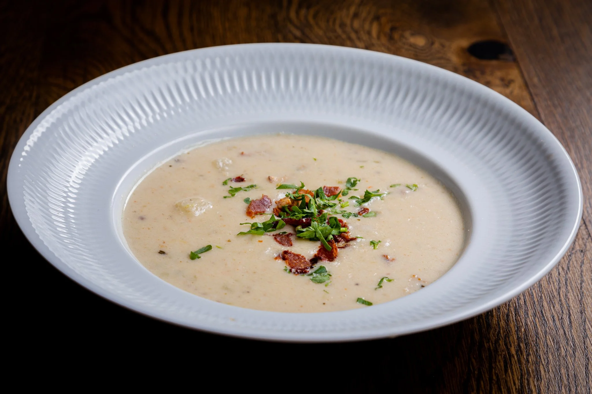 Bowl of creamy soup garnished with chopped herbs and bacon bits, placed on a dark wooden table.