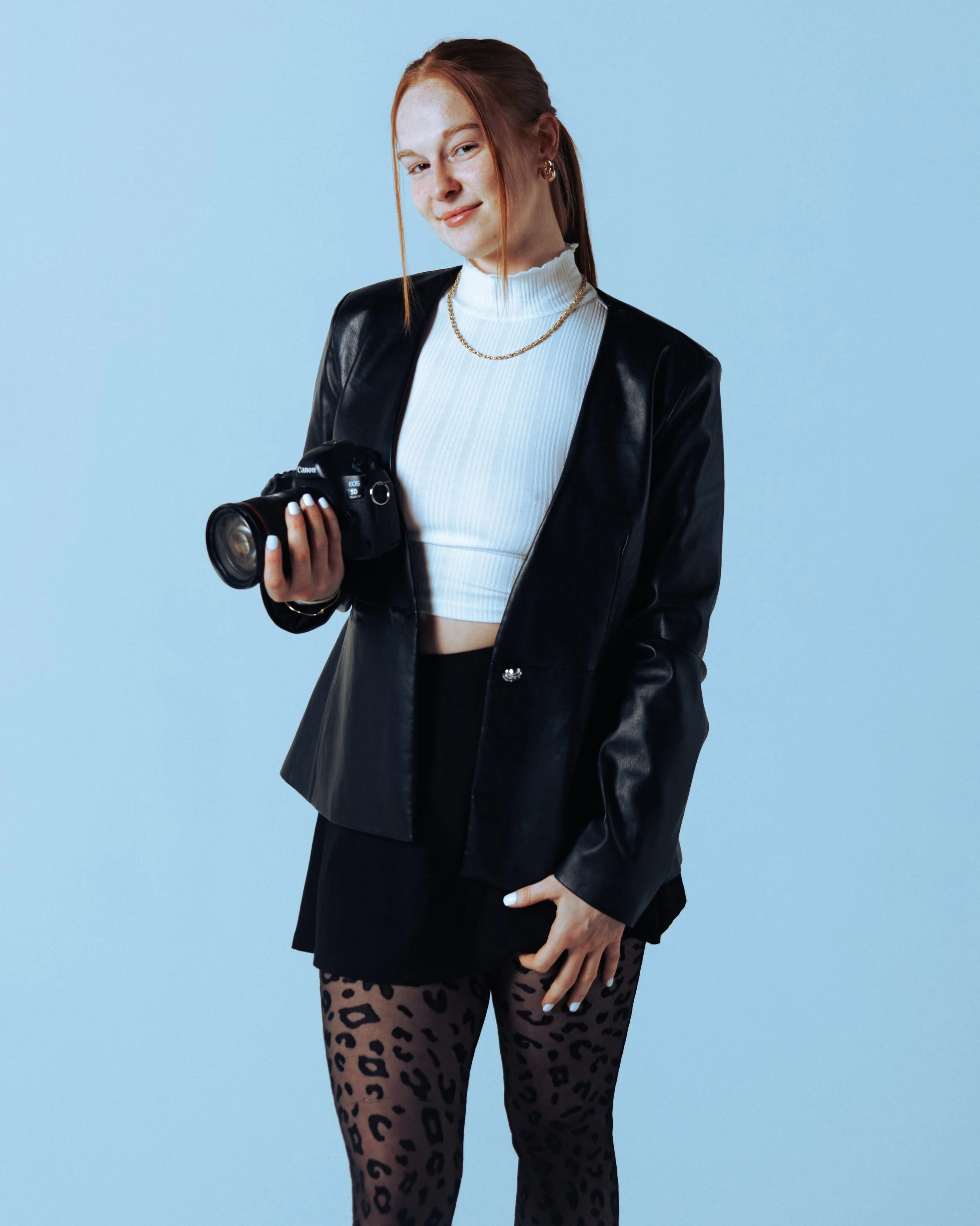 A young woman with red hair, wearing a black blazer, white cropped turtleneck, gold necklace, and leopard print leggings, holding a camera, against a light blue background.