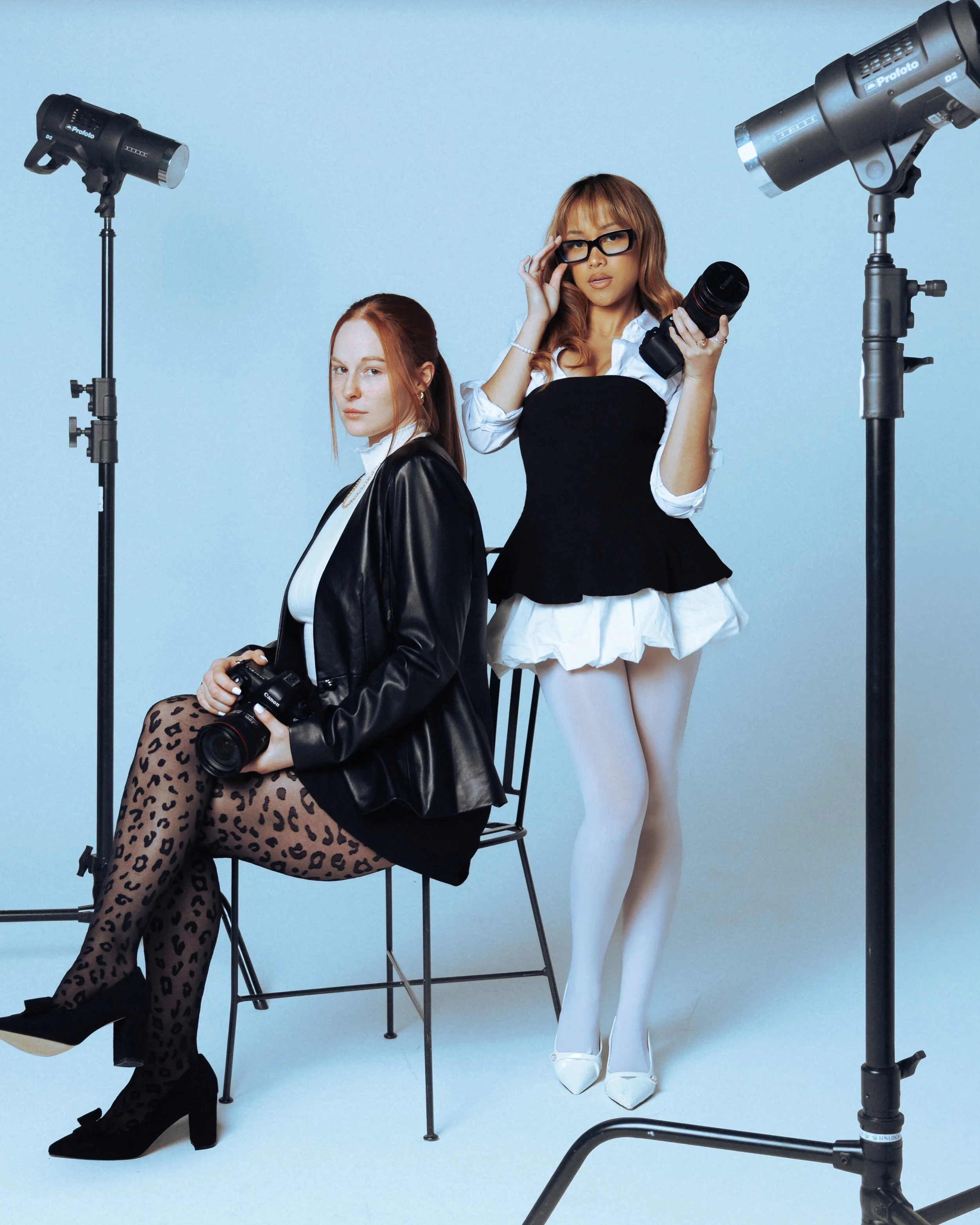 Two women in a photography studio with professional lighting. One woman is sitting on a chair, holding a camera, wearing leopard print tights, black shoes, and a black leather jacket. The other woman is standing, holding a camera lens, wearing large glasses, an outfit with a black and white top, white tights, and white high heels.