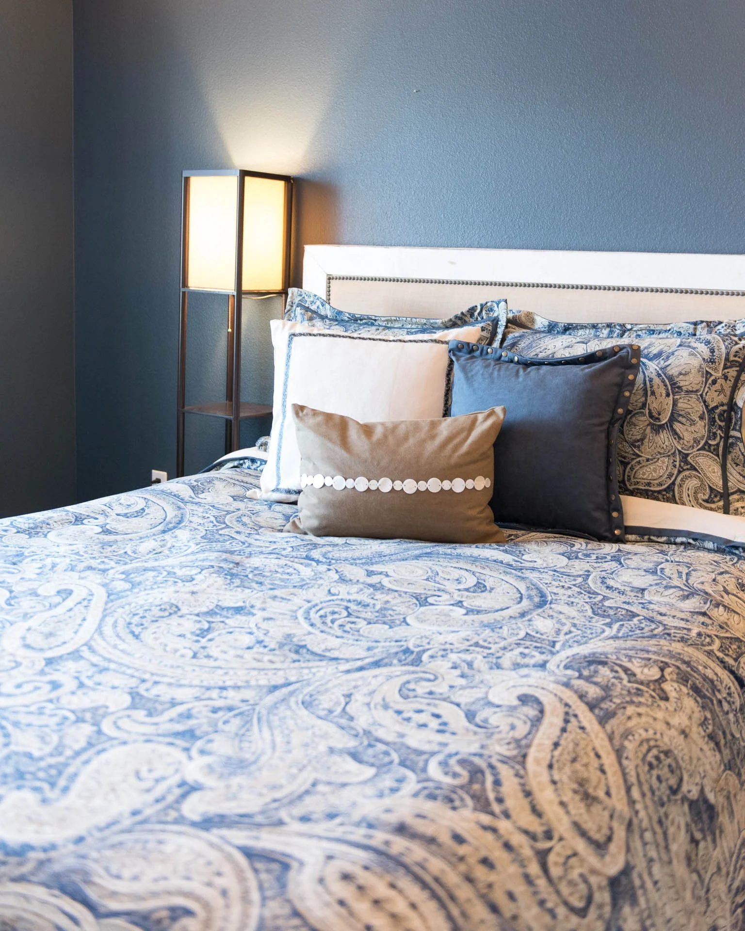 A neatly made bed with multiple decorative pillows, a floral-patterned white and blue bedspread, a white headboard, and a tall lamp on the side against a blue wall.