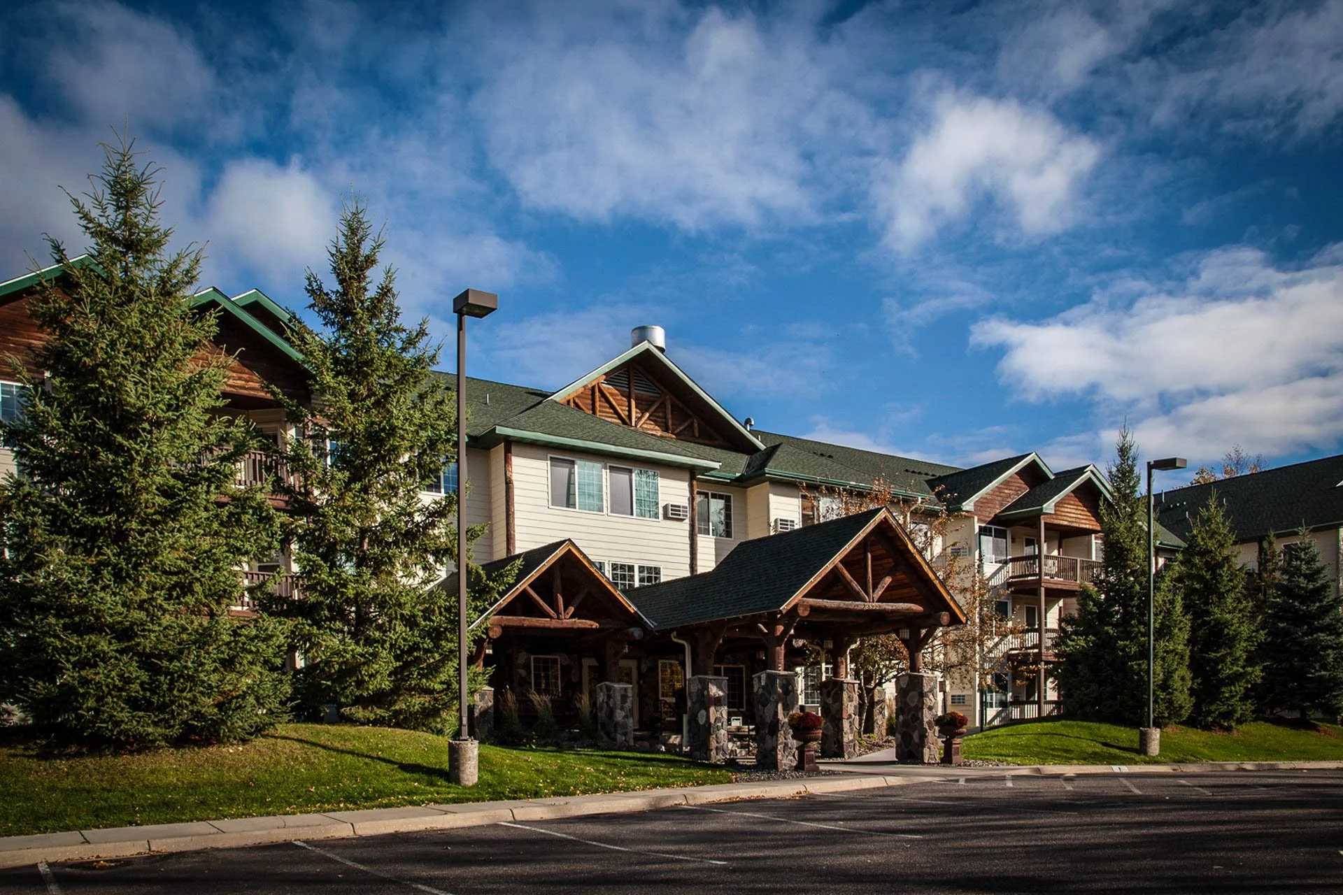 Multi-story residential building with wood accents in a suburban neighborhood, surrounded by trees, sidewalk, and street lights under a partly cloudy sky.