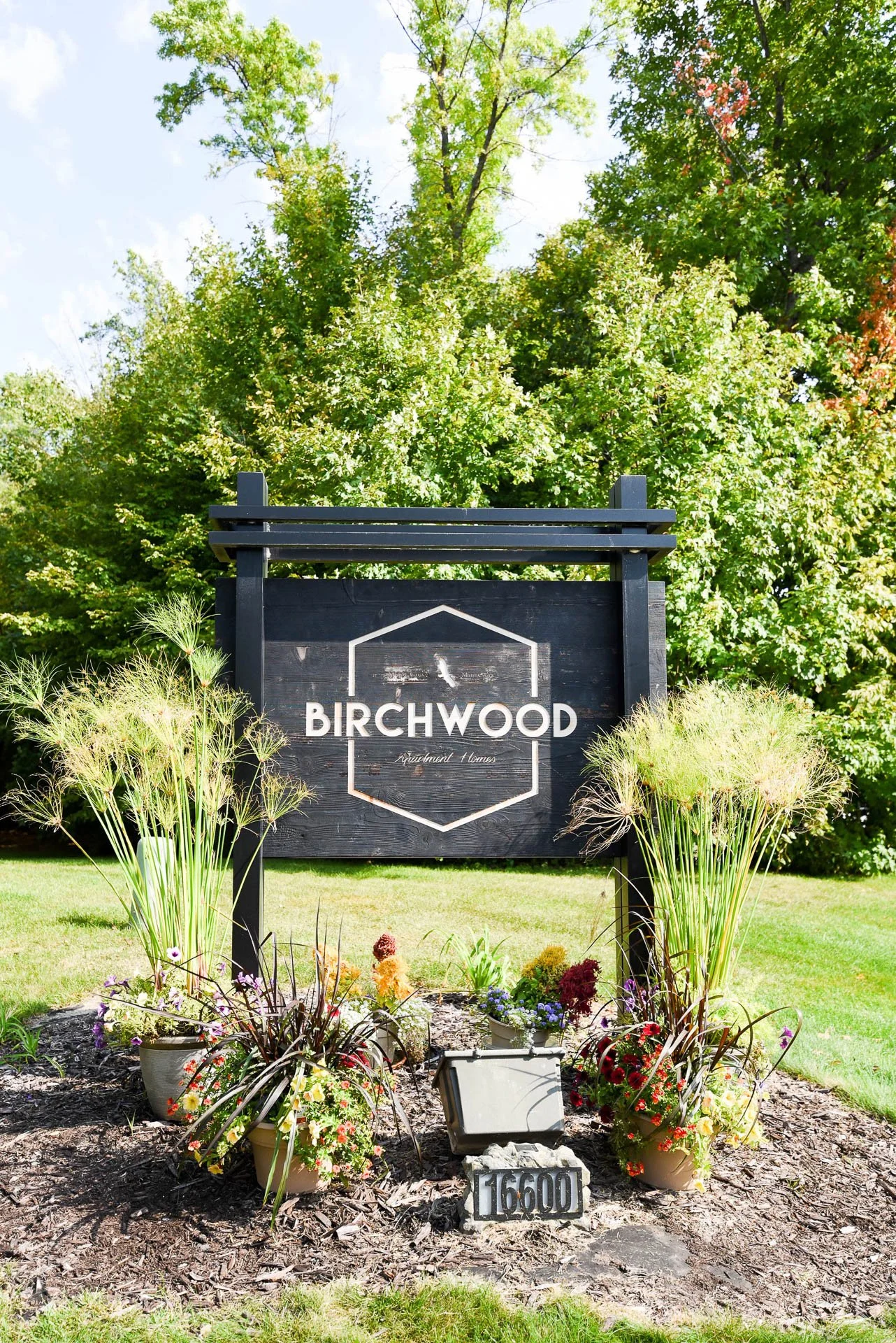 A black sign with white lettering and geometric design that reads 'Birchwood Apartment Homes,' surrounded by potted plants and flowers, with green trees in the background.