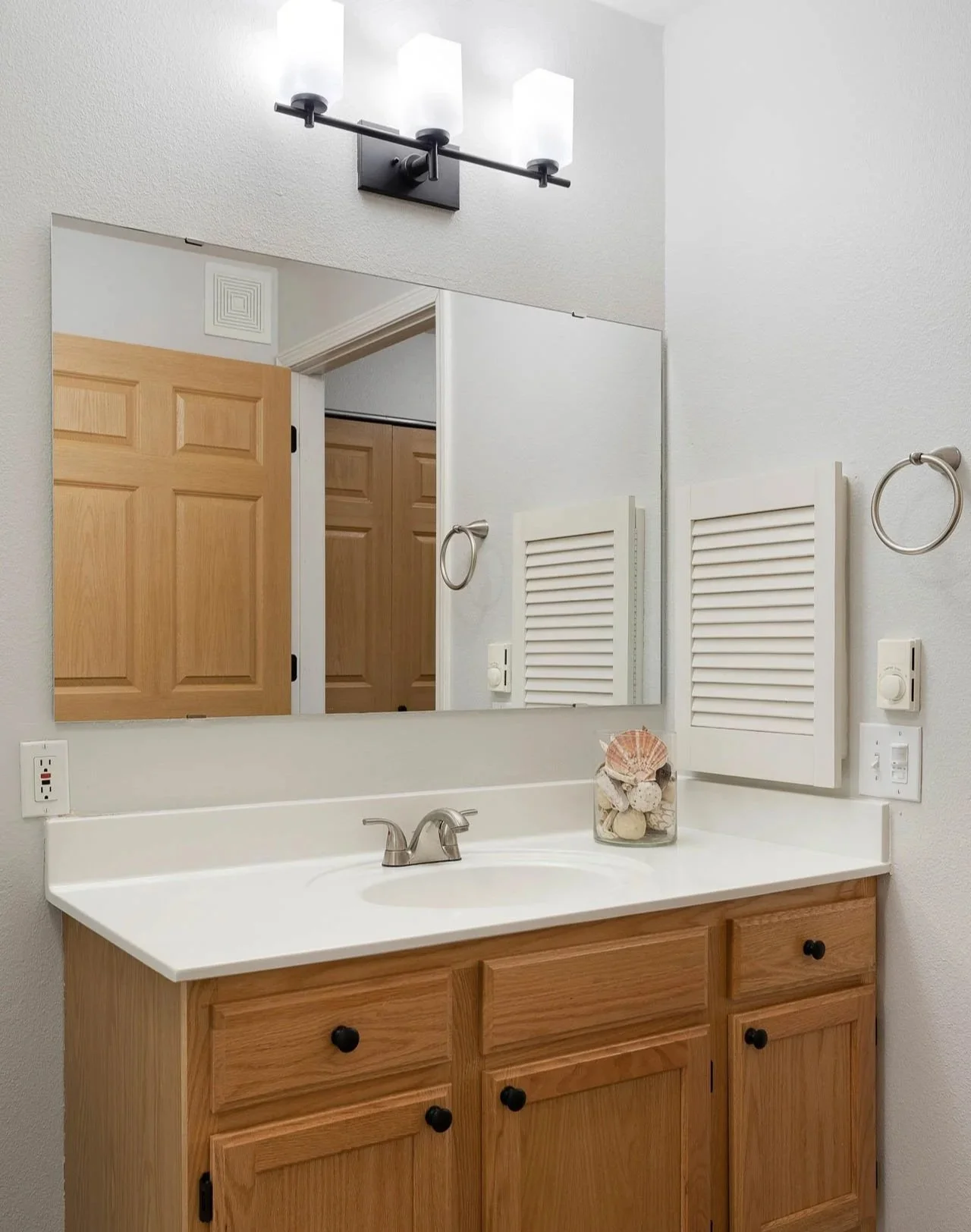 Bathroom vanity with a mirror, three light fixtures above, wooden cabinet with black knobs, shell and rock decor, and a white countertop.