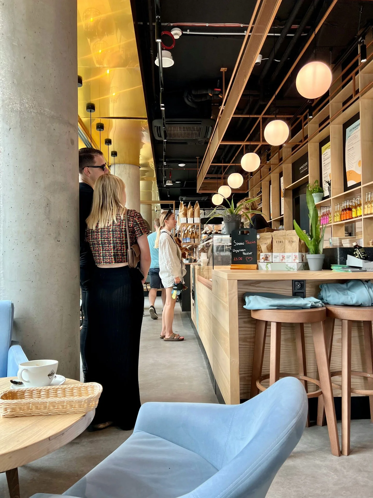 People standing in line at a modern coffee shop counter with hanging spherical lights and wooden shelving, a plant, and a chalkboard sign on the counter.