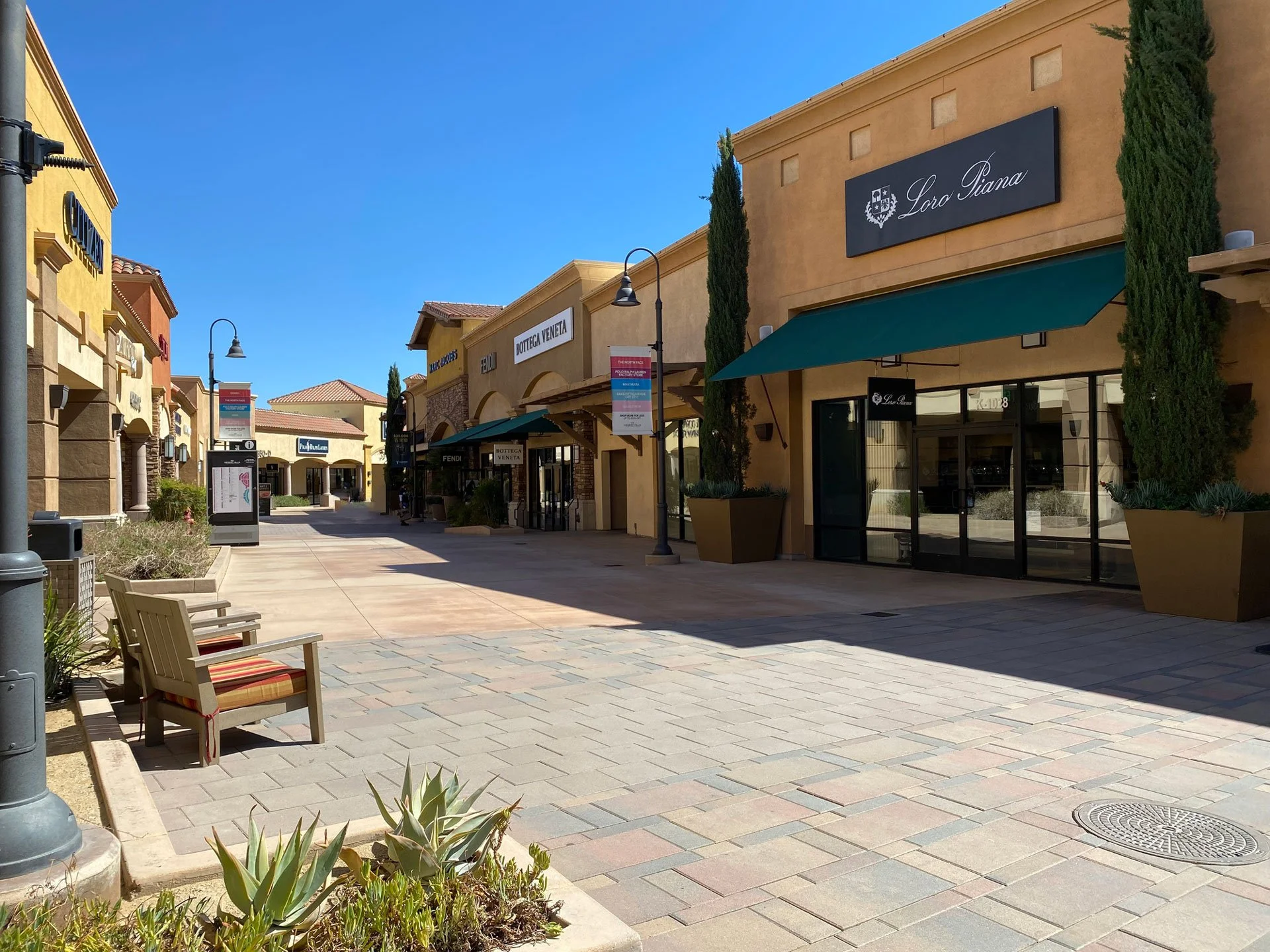 Empty shopping center outdoor walkway with storefronts. Benches and plants are visible, with a bright blue sky overhead.