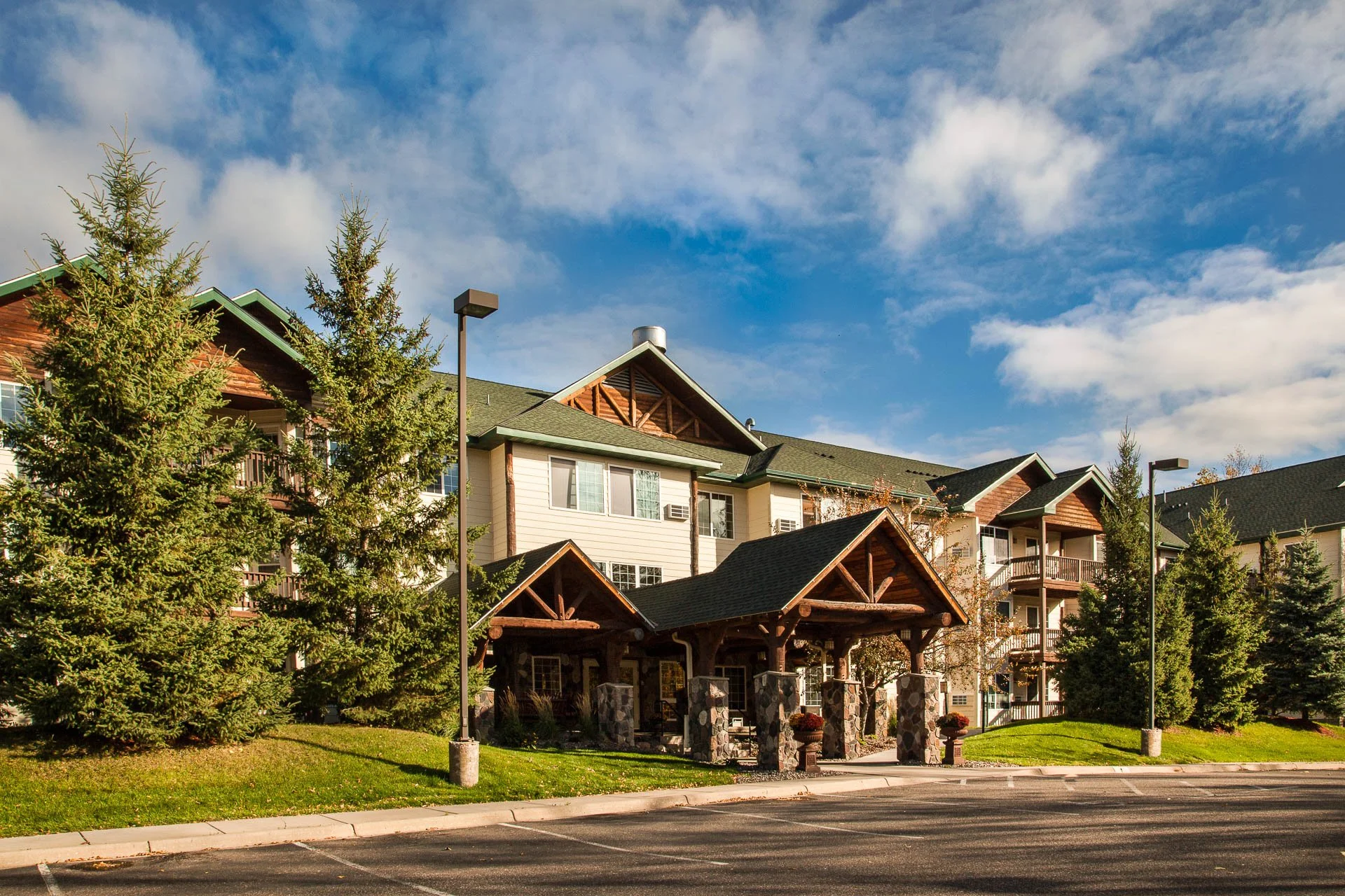 A multi-story residential apartment complex with covered entrance, surrounded by green lawns, tall evergreen trees, and street lamps under a partly cloudy blue sky.