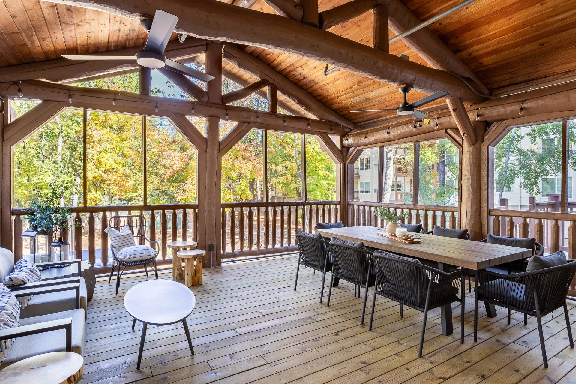 Spacious outdoor screened porch with wooden floors, ceiling, and railings, furnished with a large dining table, black chairs, a small sofa with pillows, a round coffee table, chairs, and plants, surrounded by trees with fall foliage.