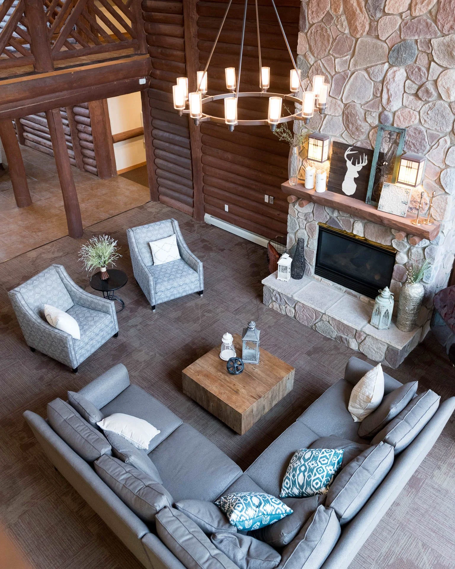Living room with a grey sectional sofa, two armchairs, a wooden coffee table, a stone fireplace, and a chandelier with multiple candles hanging from the ceiling in a log cabin style interior.
