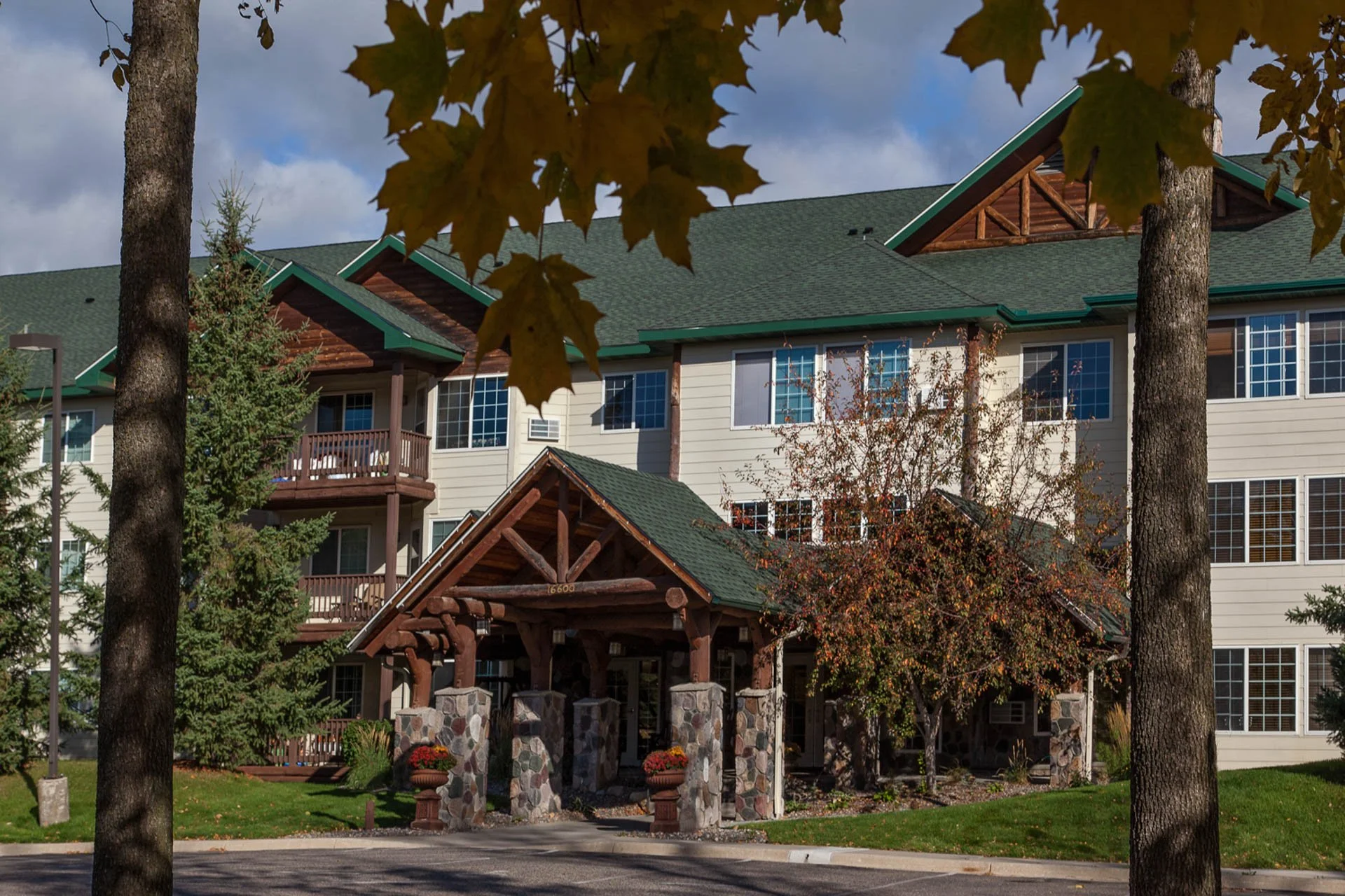 Exterior view of a multi-story residential building with a green roof, surrounded by trees with autumn leaves, and a stone and wood entrance structure.