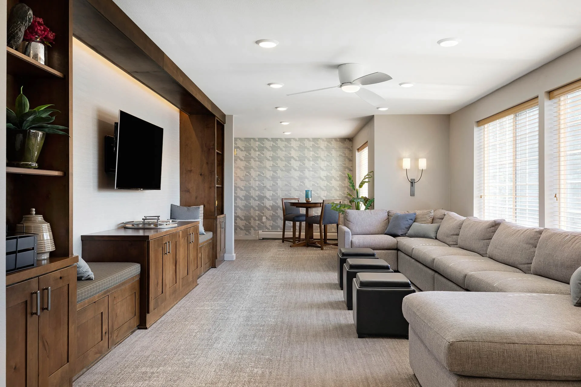 Living room with beige sectional sofa, black ottomans, wall-mounted TV, wooden entertainment unit with shelving, wall sconce, ceiling fan, large windows with blinds, and a small dining area with a round table and chairs in the background.