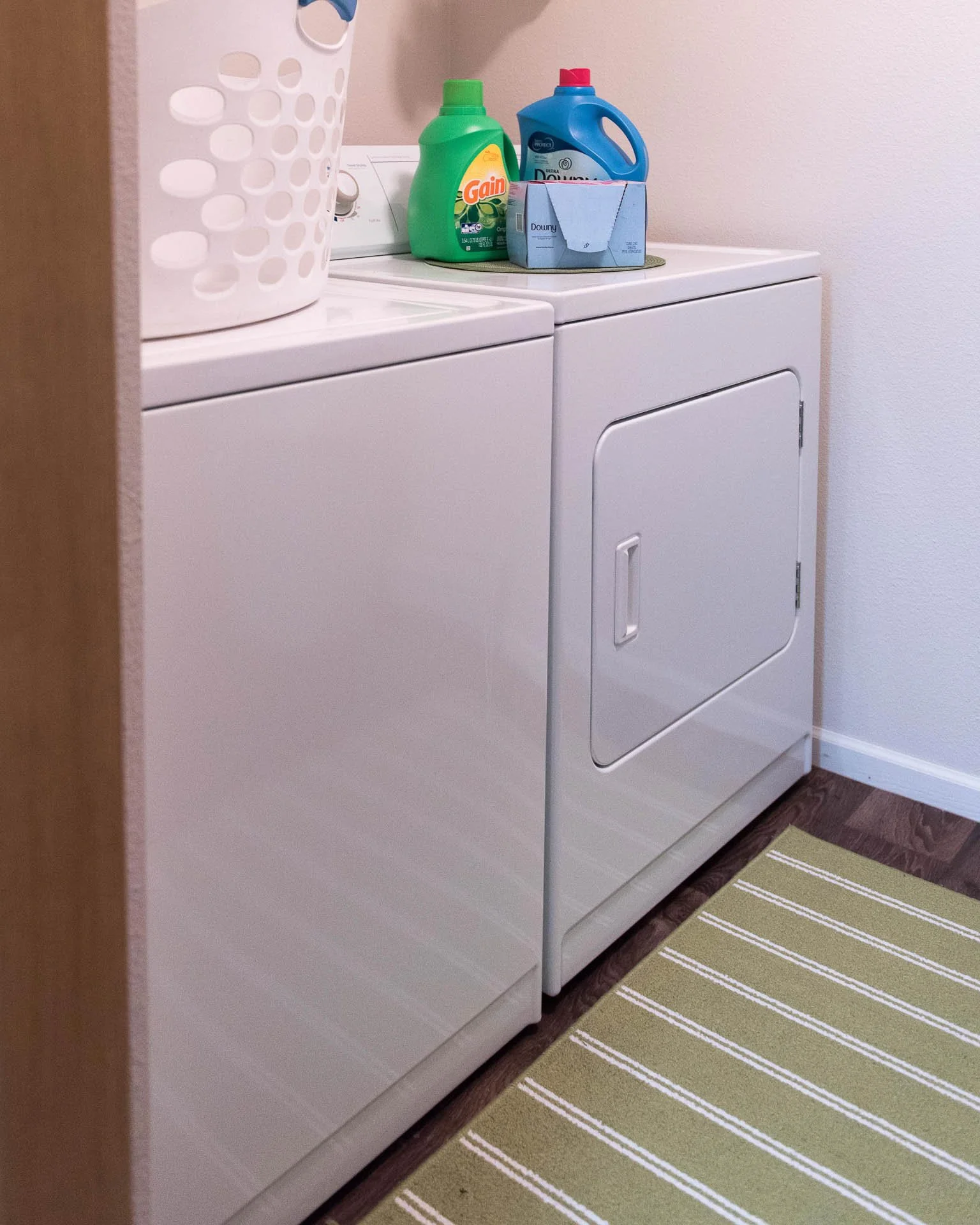 A laundry room with a white washing machine and dryer side by side. On top of the appliances are laundry detergents and a box of soap. There's a white laundry basket on the washing machine and a green and white striped rug on the wooden floor.