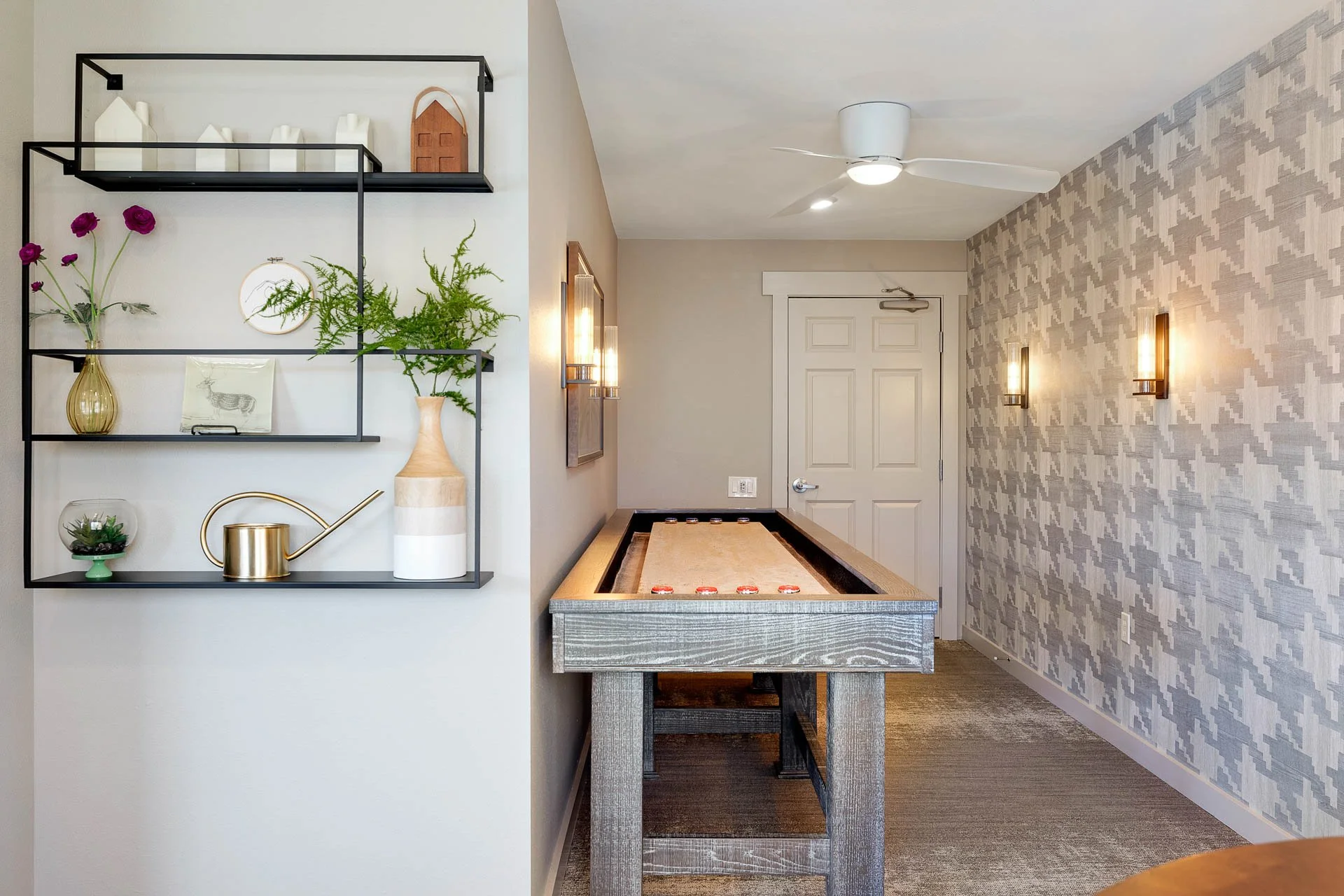 Indoor game room featuring a shuffleboard table and a decorative wall with black shelving holding vases, plants, and decorative items, with wall-mounted lights and patterned wallpaper.