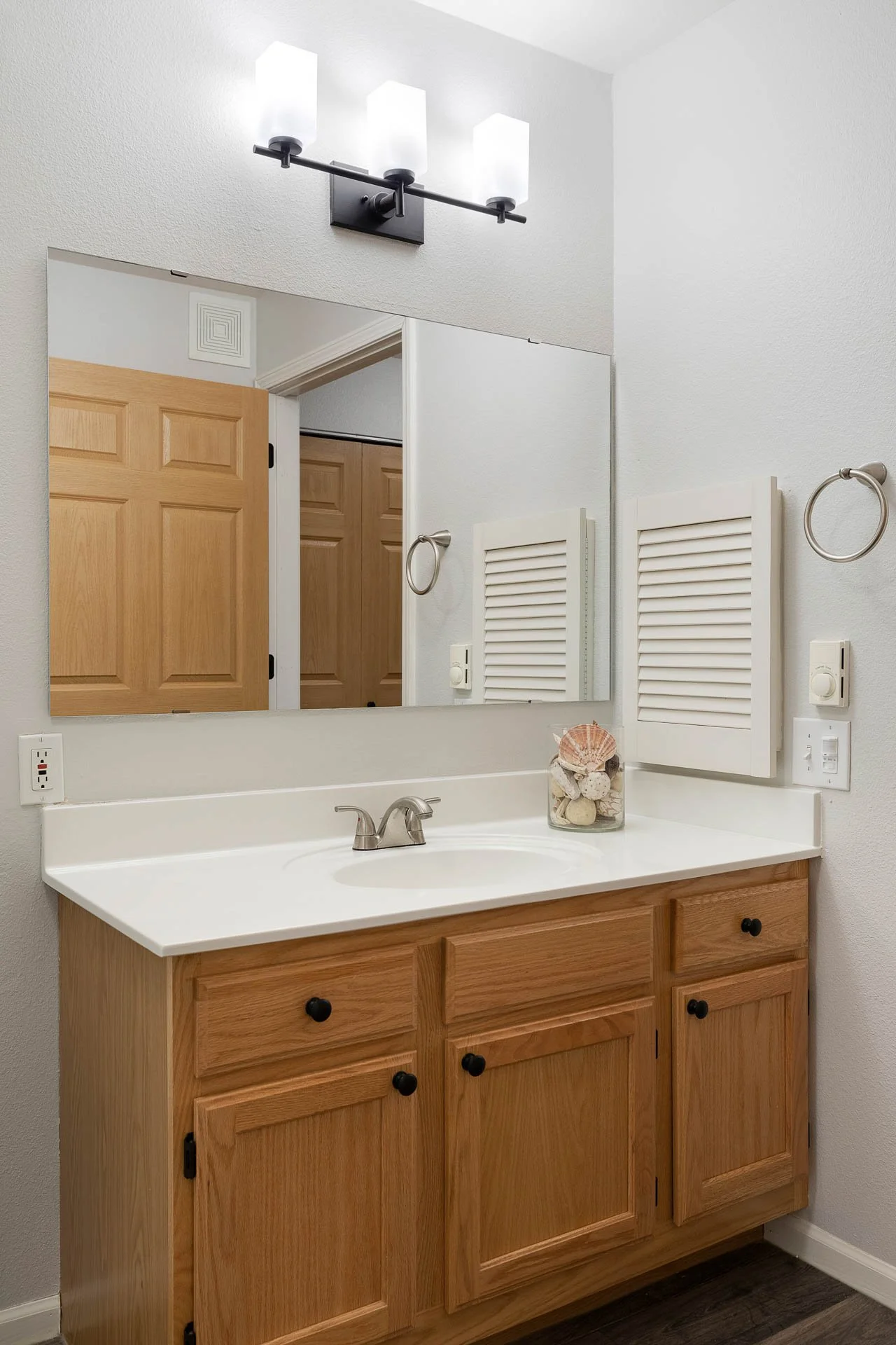 Bathroom vanity with a mirror, three light fixtures above, wooden cabinet with black knobs, shell and rock decor, and a white countertop.