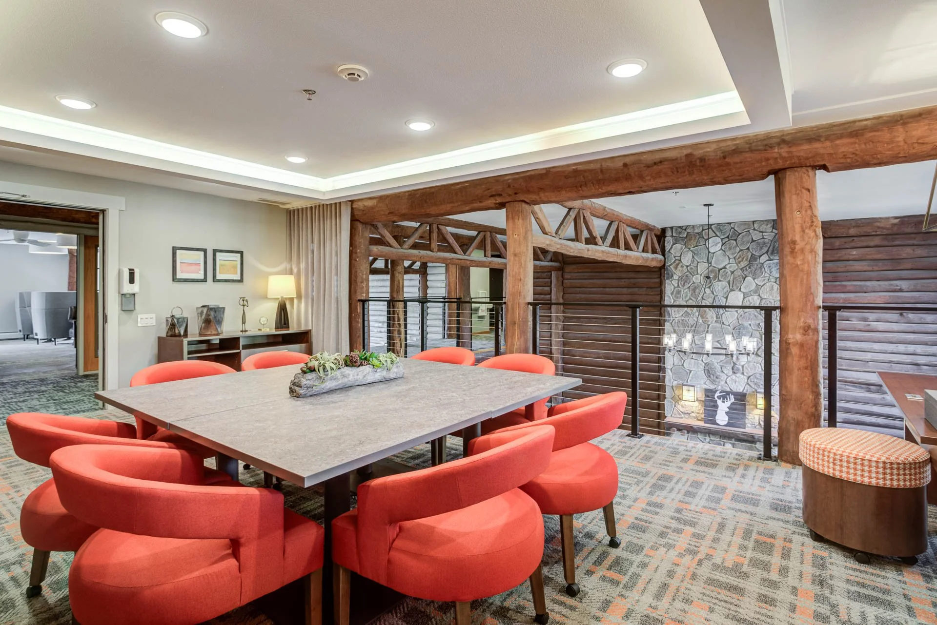 Interior of a rustic-style dining or meeting area with a large rectangular table surrounded by eight coral-colored chairs, wood and stone accents, a stone fireplace, and decorative lighting.