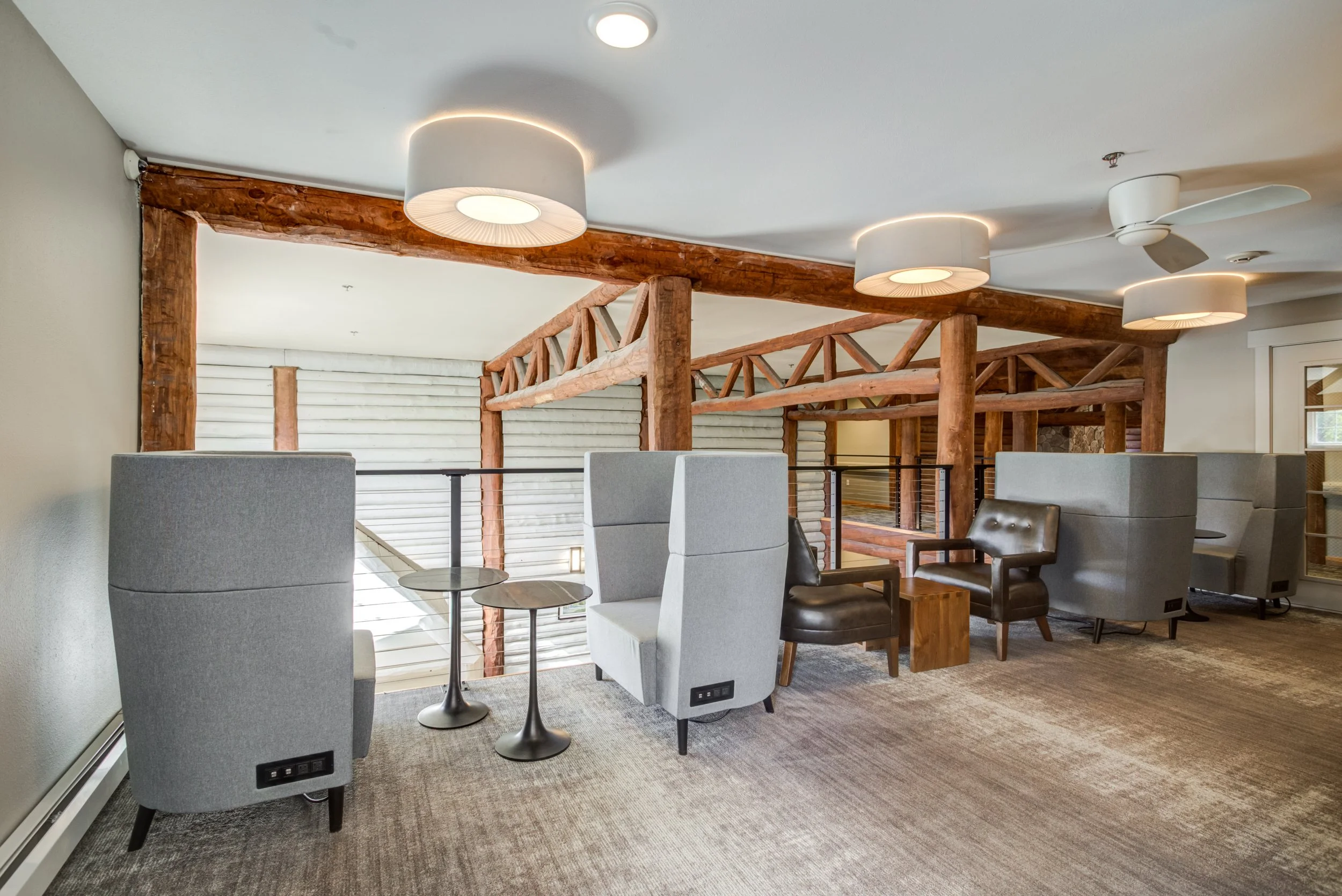 Interior view of a modern open-space lounge area with gray high-back chairs, small round tables, wooden beams, and ceiling lights.