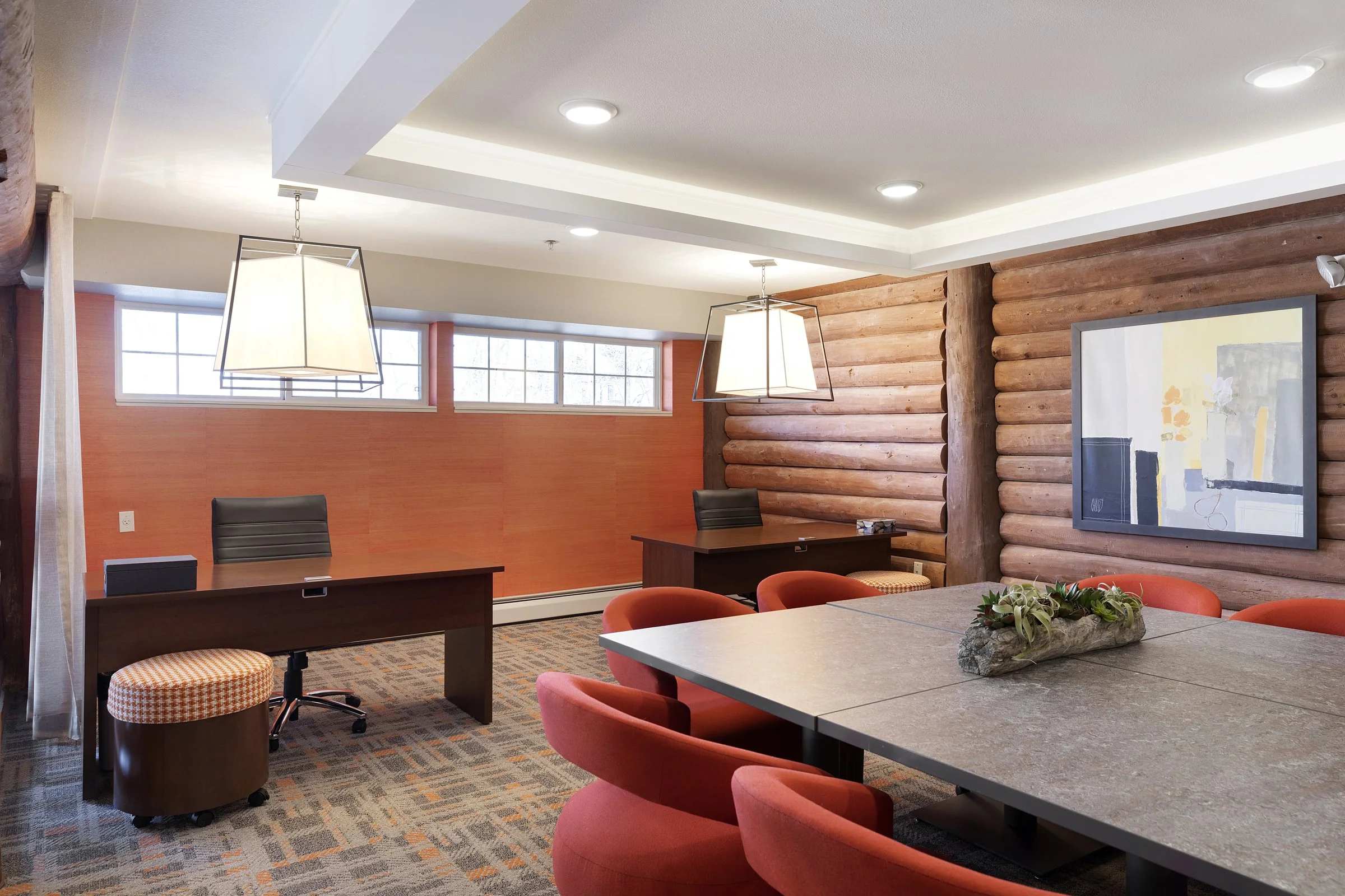 Modern conference room with wood log walls, large window, rectangular table with red chairs, and two desks with black chairs.