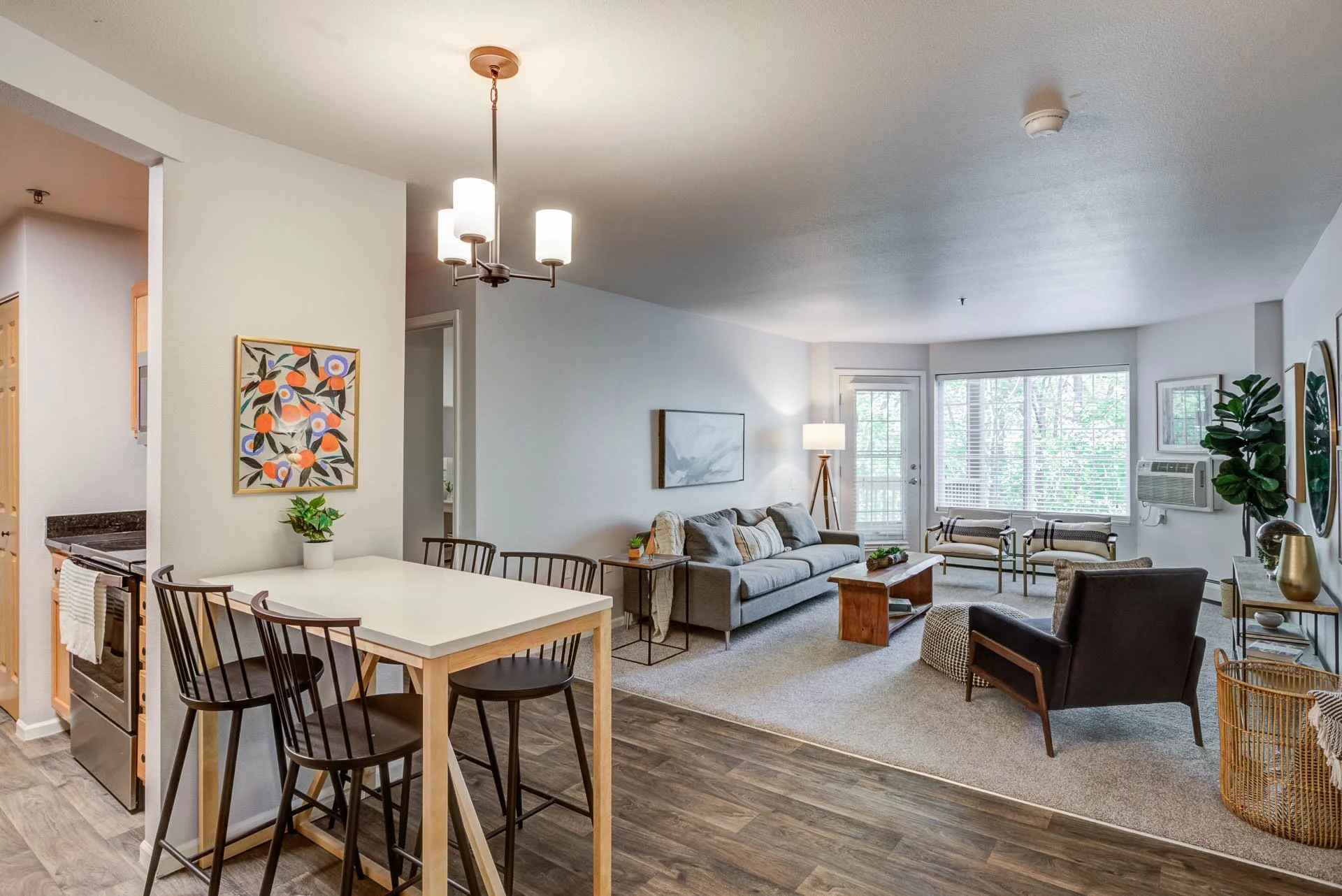 A modern open-concept living room and dining area with large windows, a gray sofa, black armchair, wooden coffee table, indoor plants, and artwork.