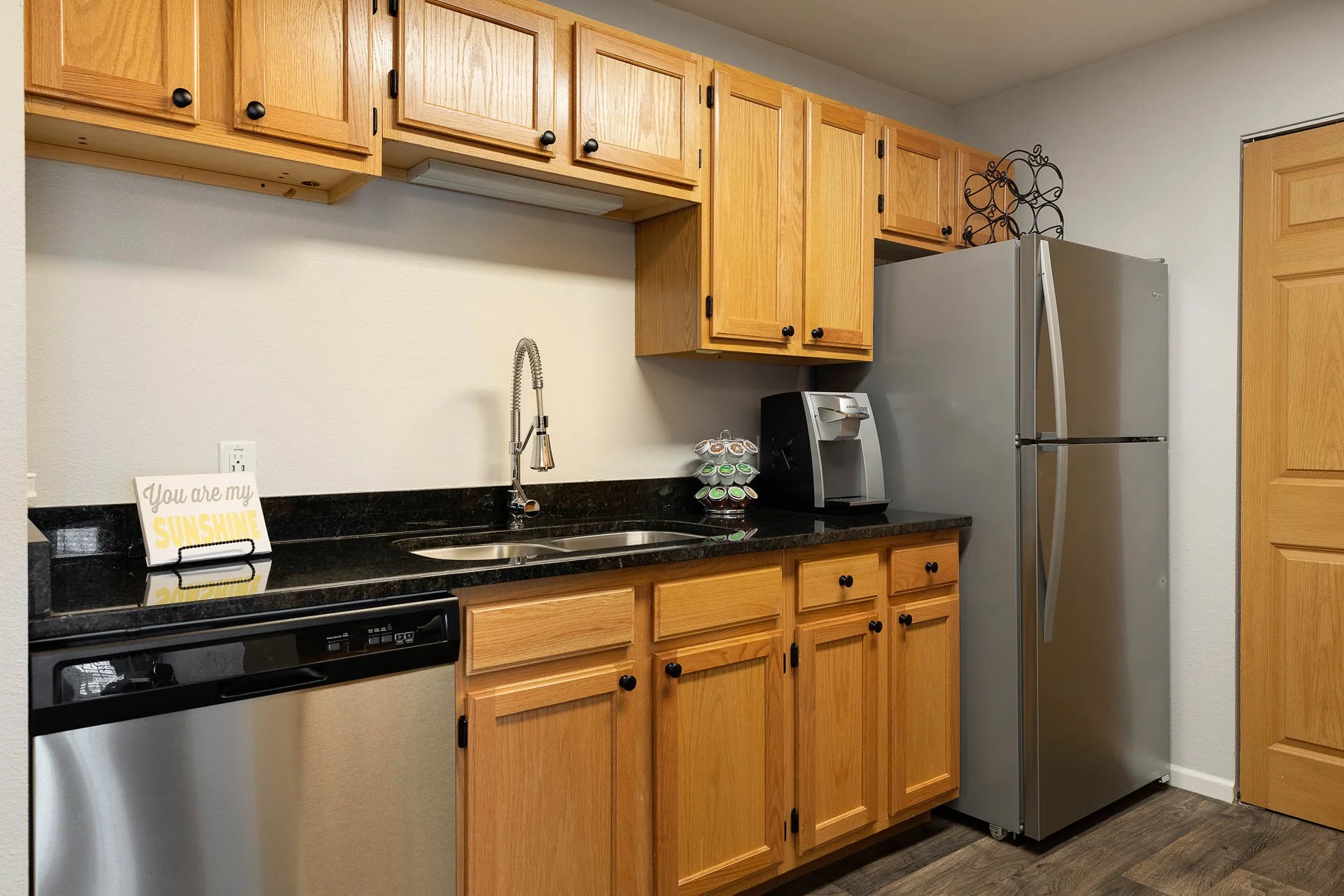 Kitchen with light wooden cabinets, black countertops, stainless steel fridge and dishwasher, coffee maker, and a decorative sign that says 'You are my sunshine' near the sink.