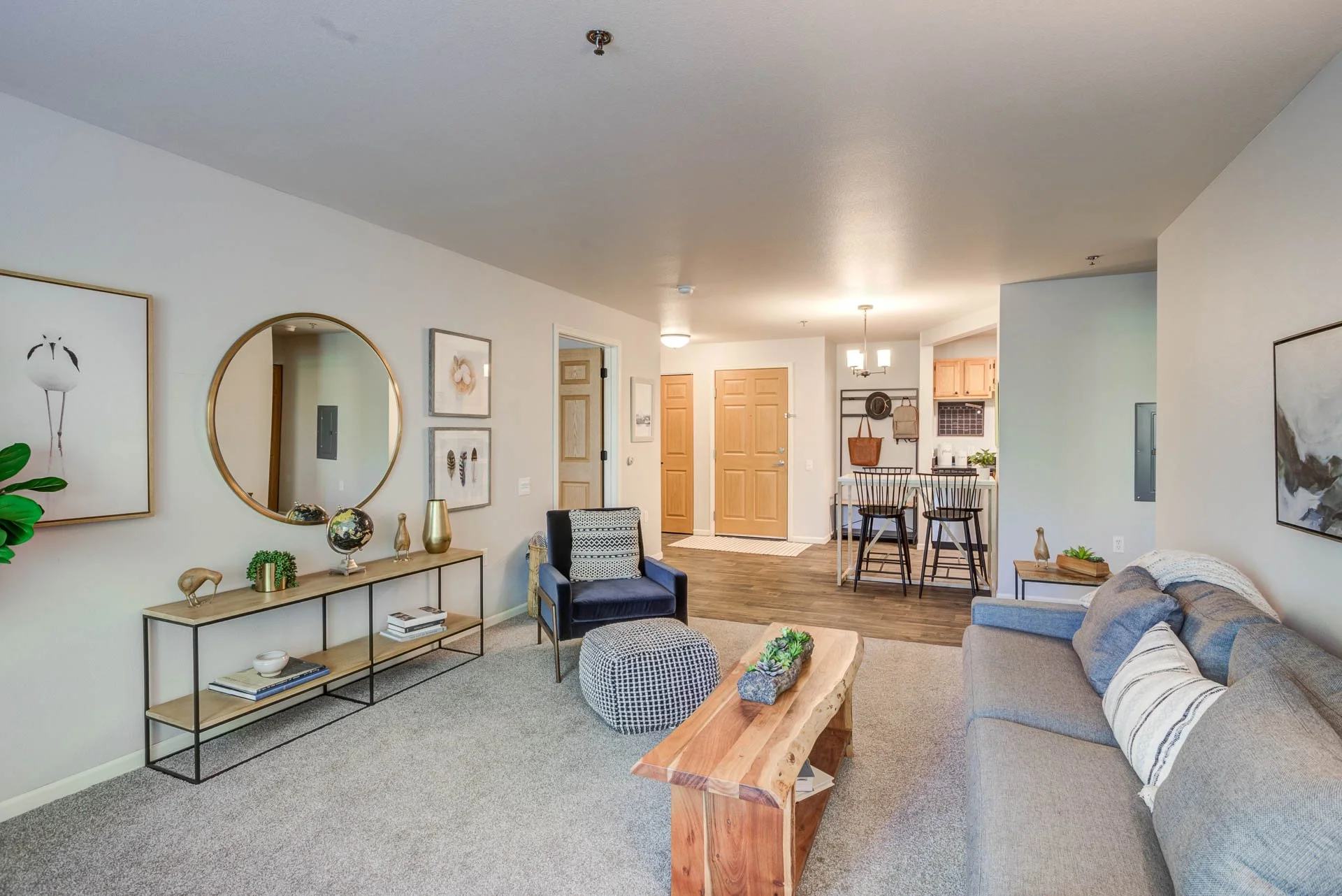 Living room with gray sofa, black armchair, beige area rug, wooden coffee table, wall art, mirror, and decor items, leading to a dining area and kitchen with wooden cabinets and barstools.