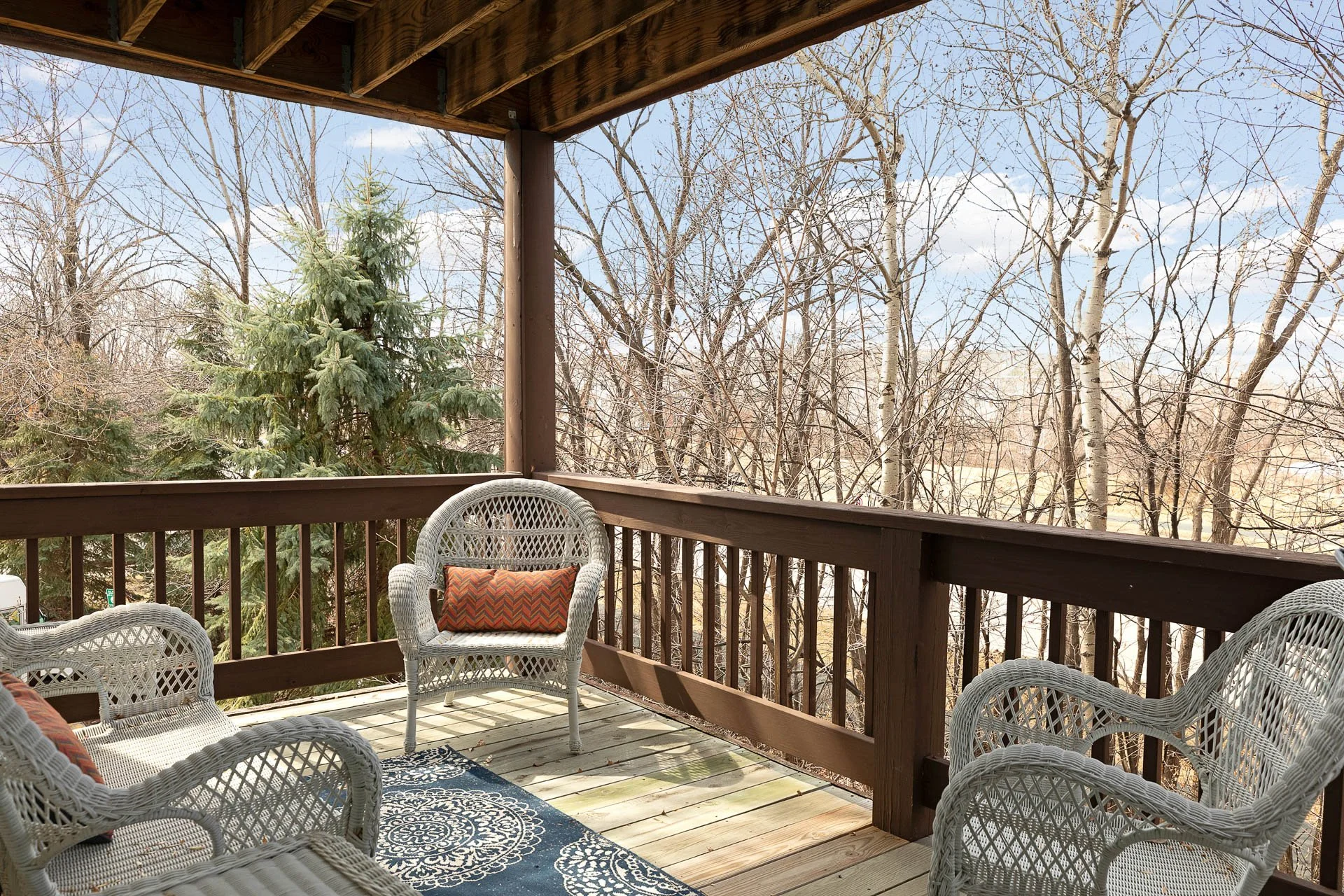 View from a wooden balcony with white wicker chairs and a cushion, overlooking leafless trees under a partly cloudy sky.