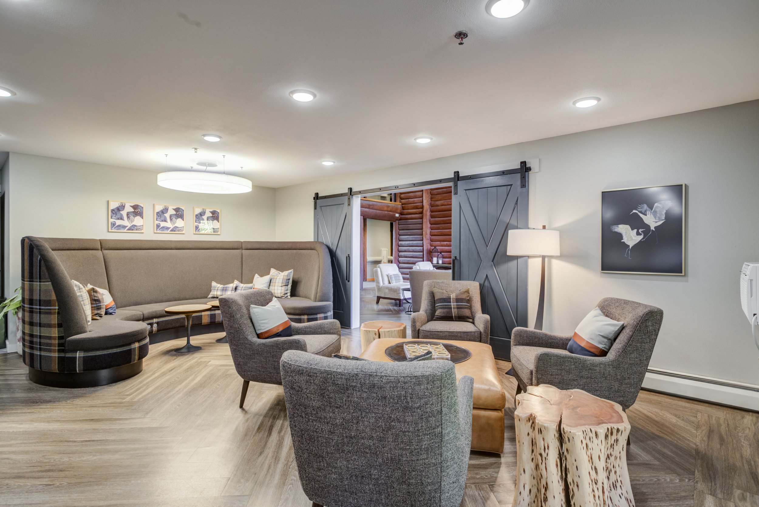 A cozy lounge area with six upholstered armchairs arranged around a wooden coffee table, a large beige curved sofa with plaid pillows, standing lamp, wall art of birds, and black barn doors leading to another room with log walls.