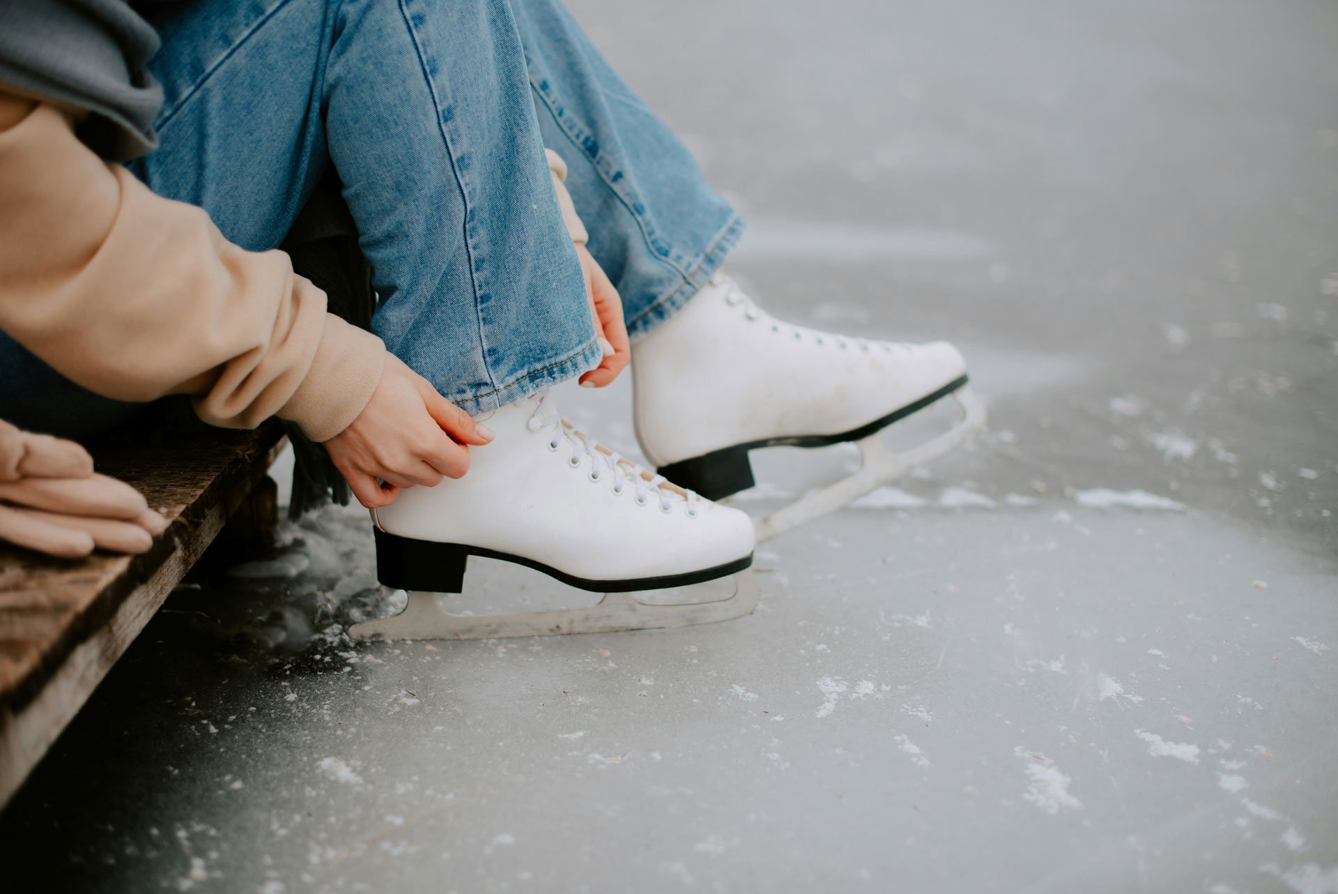 Person in blue jeans and white ice skates sitting on a bench on an ice rink, tying their skates.