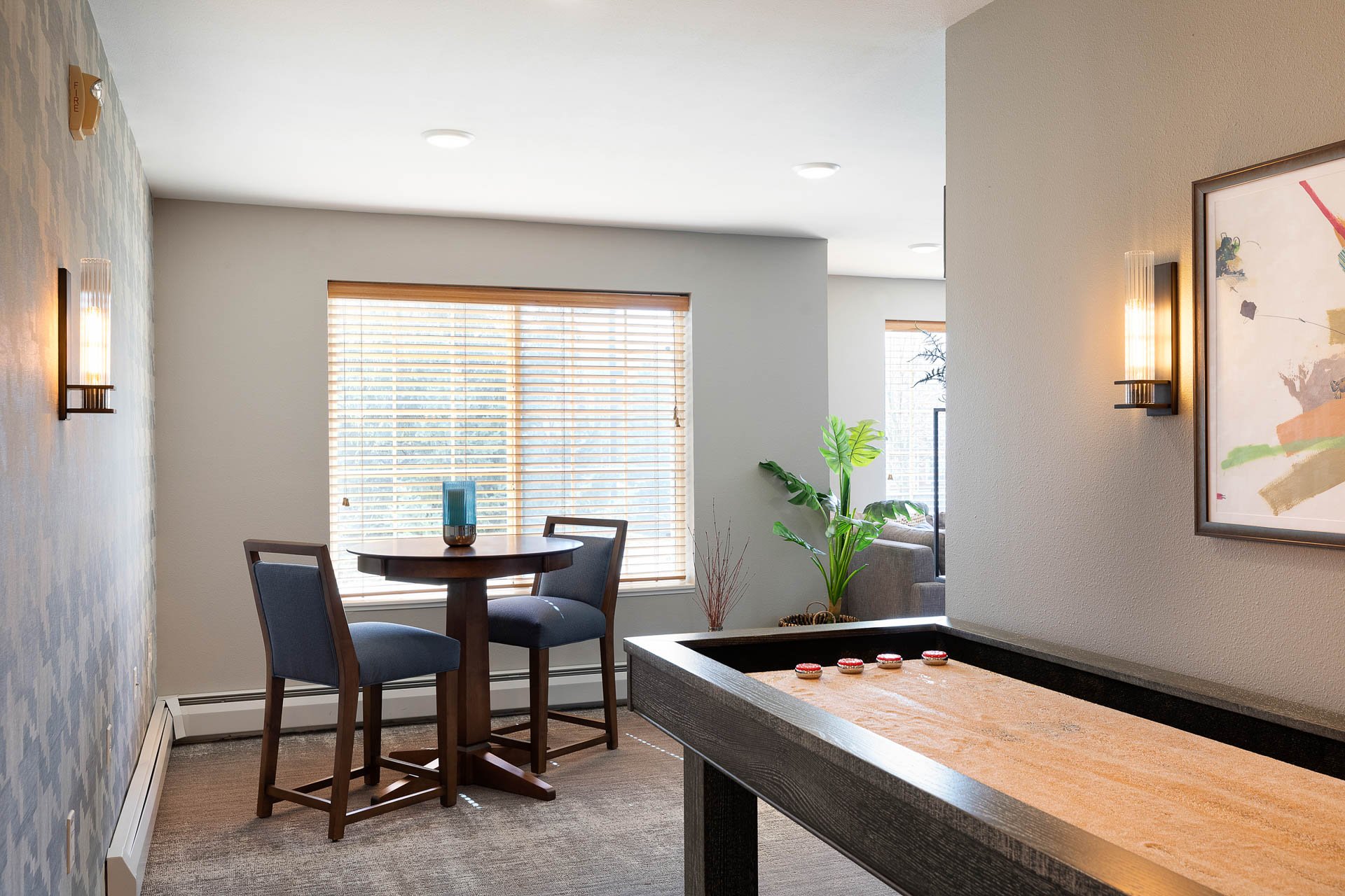 Interior of a living space with two wall-mounted lamps, a framed colorful artwork, a small round dining table with two chairs, a large window with blinds, a potted plant, and a shuffleboard table.