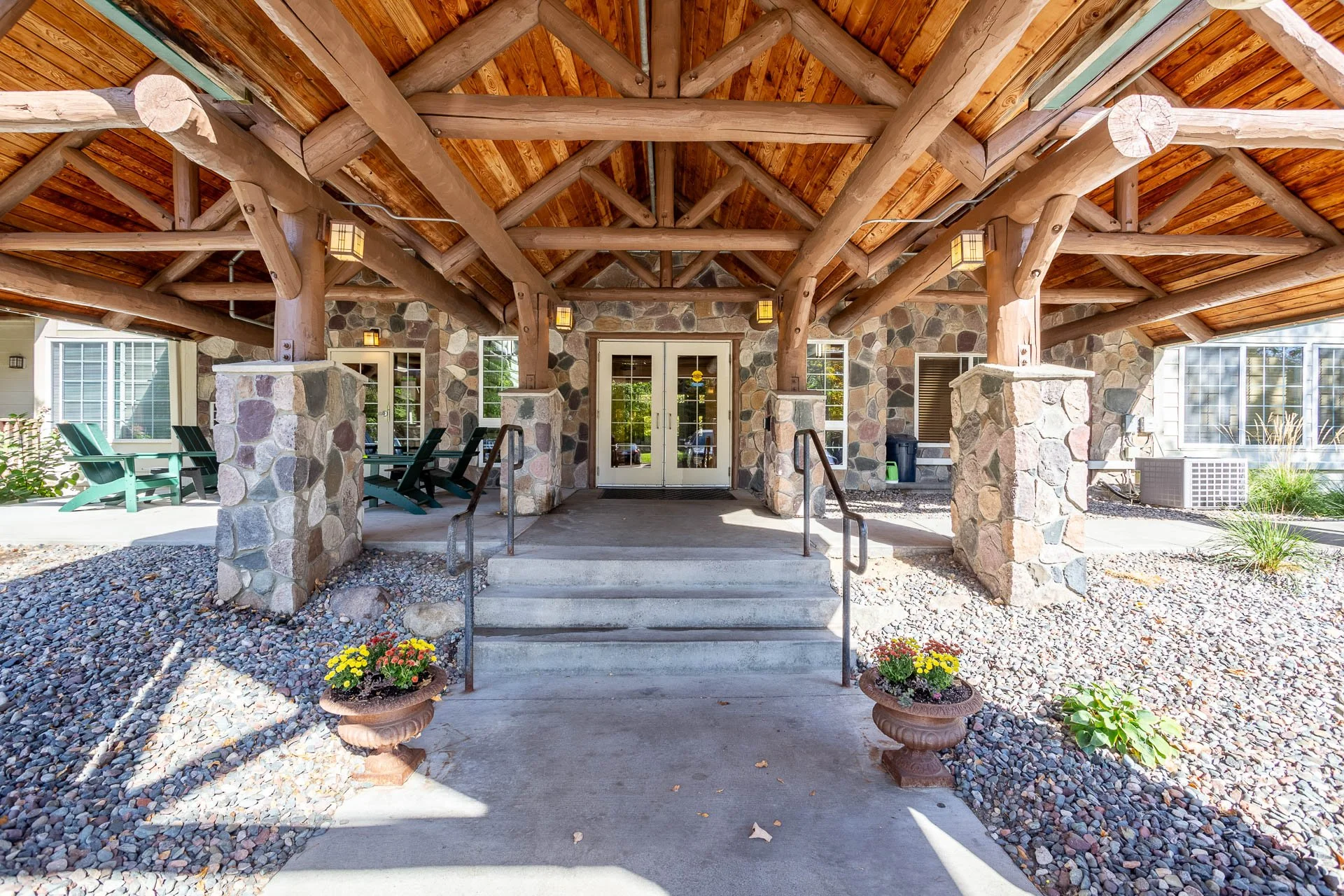 Covered apartment porch area with stone columns, wooden beams, and steps leading to glass double doors, with potted flowers and outdoor seating visible.