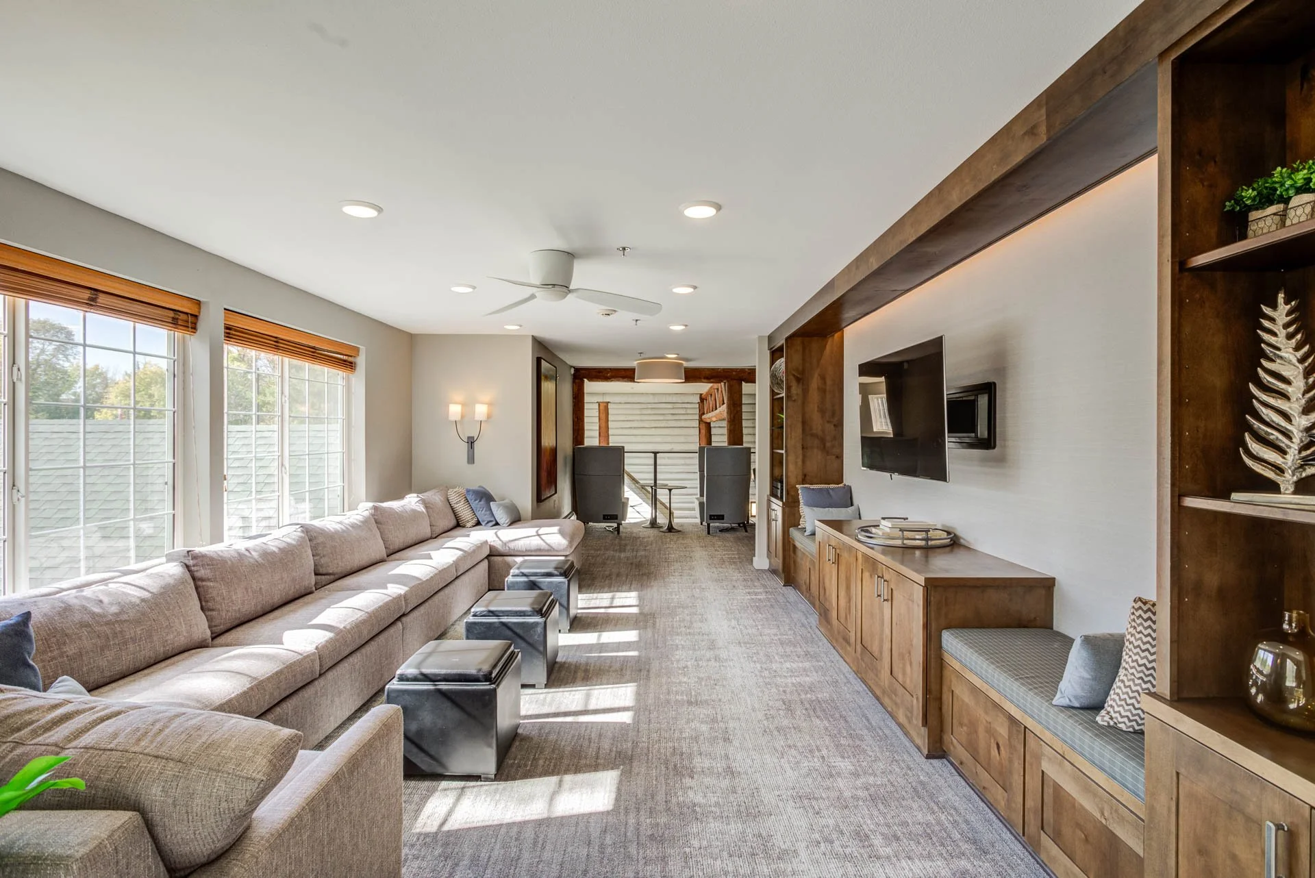 Living room with large beige sectional sofa, multiple windows with wooden blinds, a wall-mounted TV, built-in wooden shelving, and a view of stairs leading to a lower level.