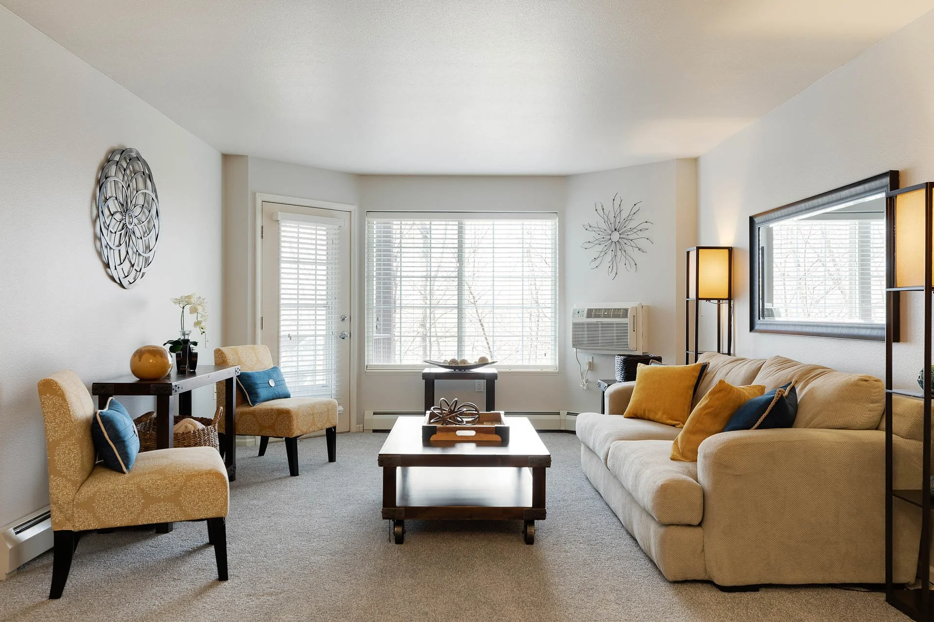 Living room with beige sofa, blue and yellow pillows, coffee table, two matching armchairs, wall art, large window with blinds, floor lamp, wall-mounted air conditioner, and decorative accents.