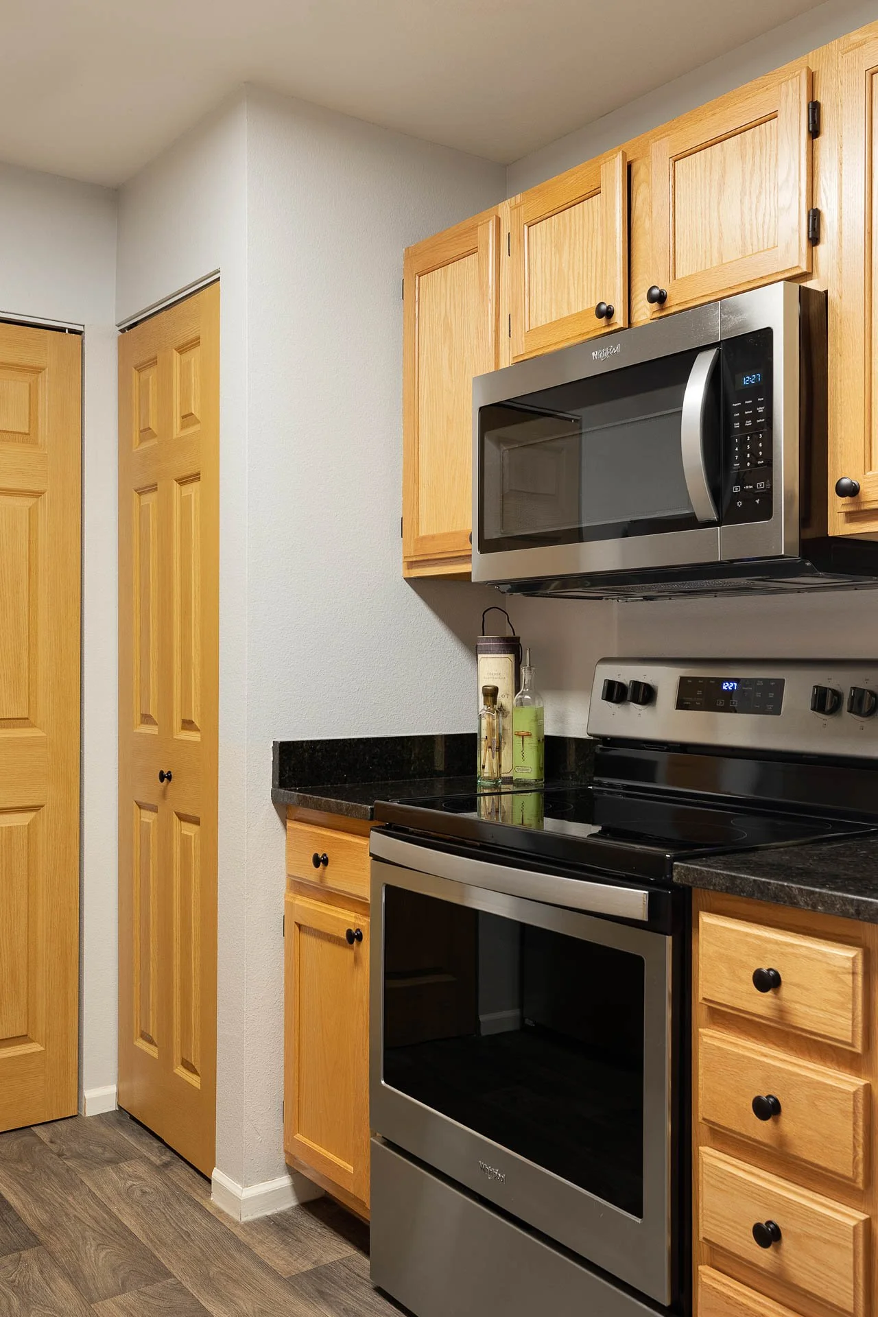 Kitchen with light wood cabinets, a black countertop, stainless steel microwave above an oven, and a bottle of oil and green soap on the counter.