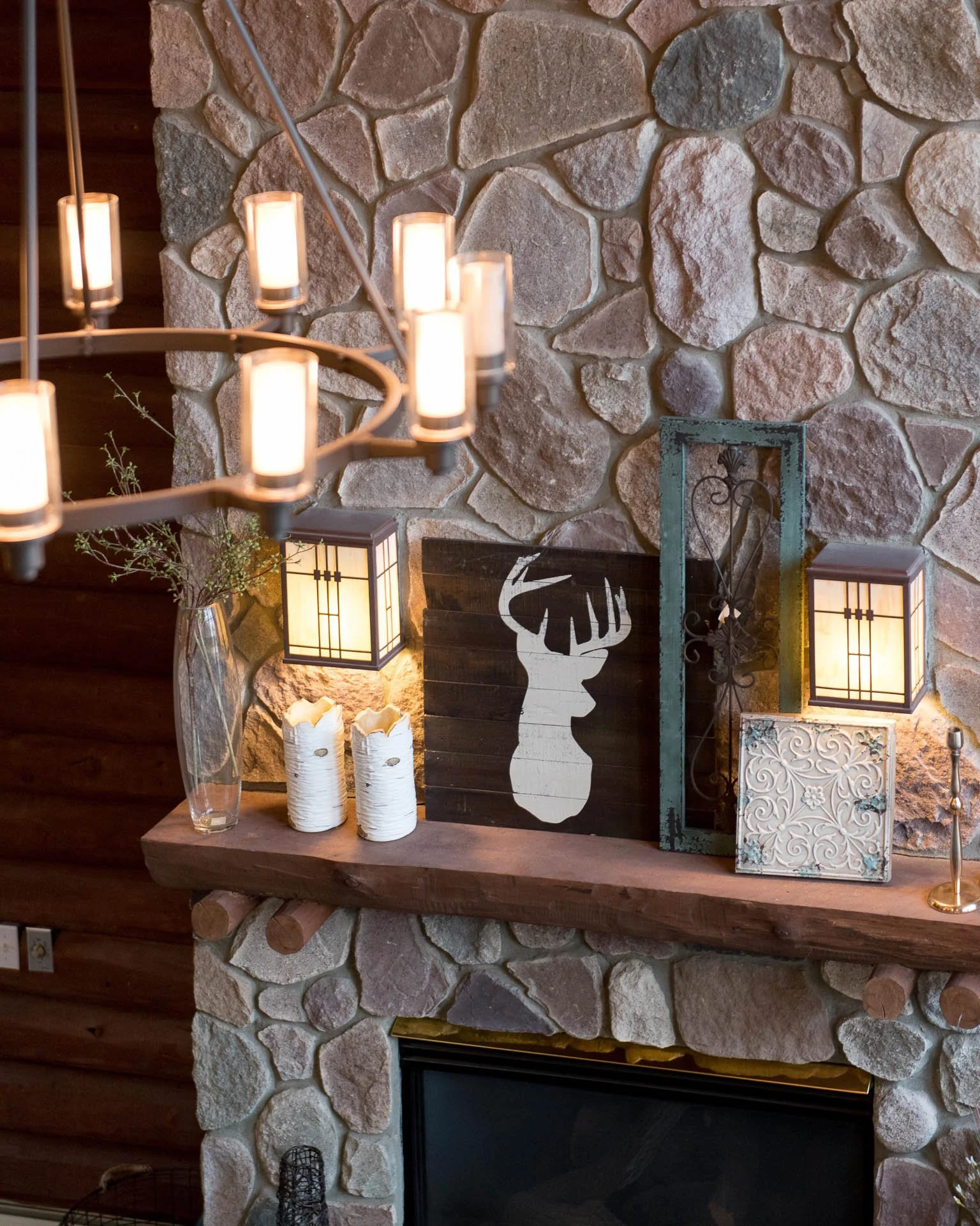 Decorative fireplace mantel with candle light fixtures, a silhouette of a deer head, a lantern, a glass vase with greenery, two white candles, and ornamental tiles set against a stone wall.