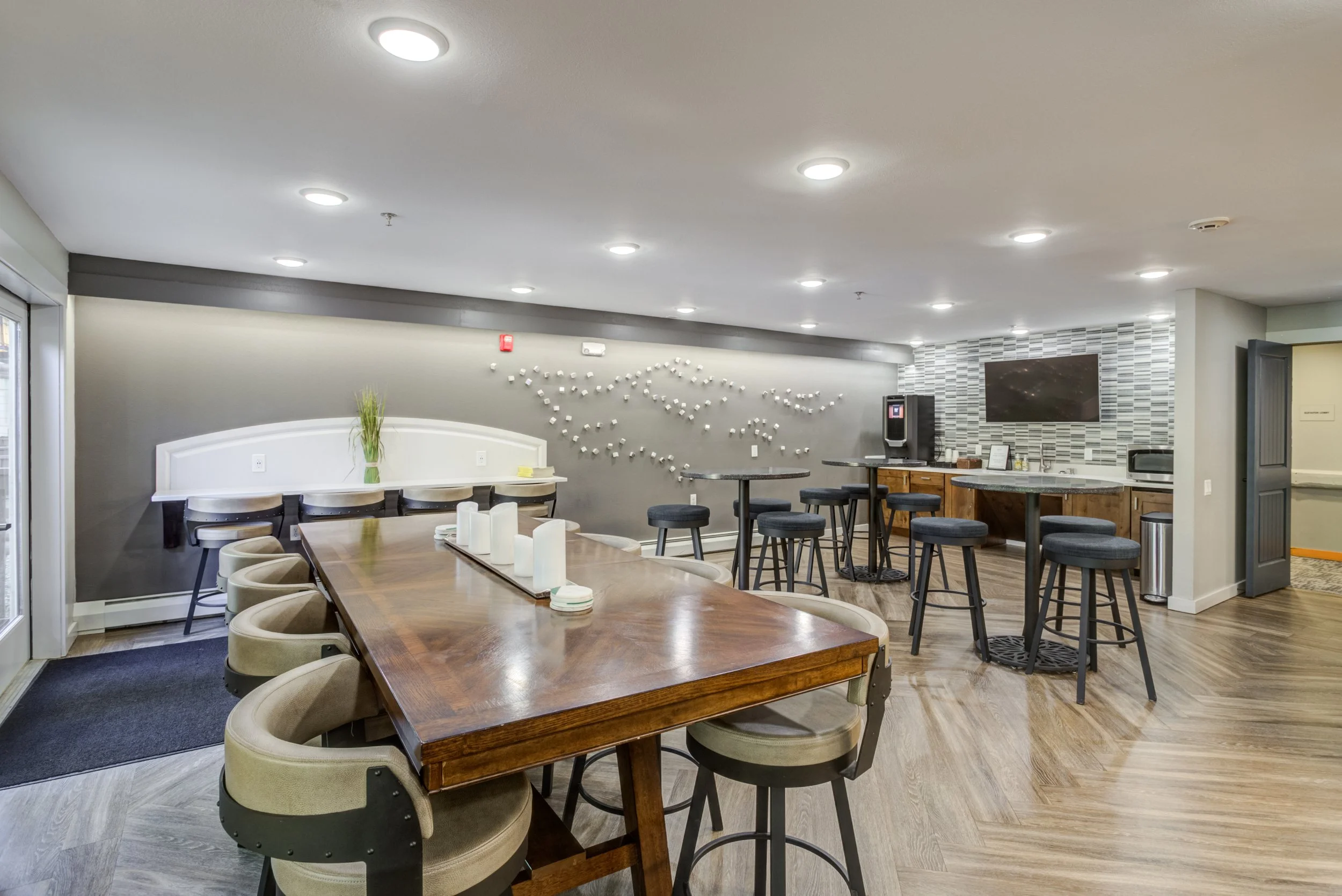 Interior of a modern lounge or dining area with a large wooden table, beige chairs, high stools, a wall-mounted TV, and a small bar area with a countertop and appliances.