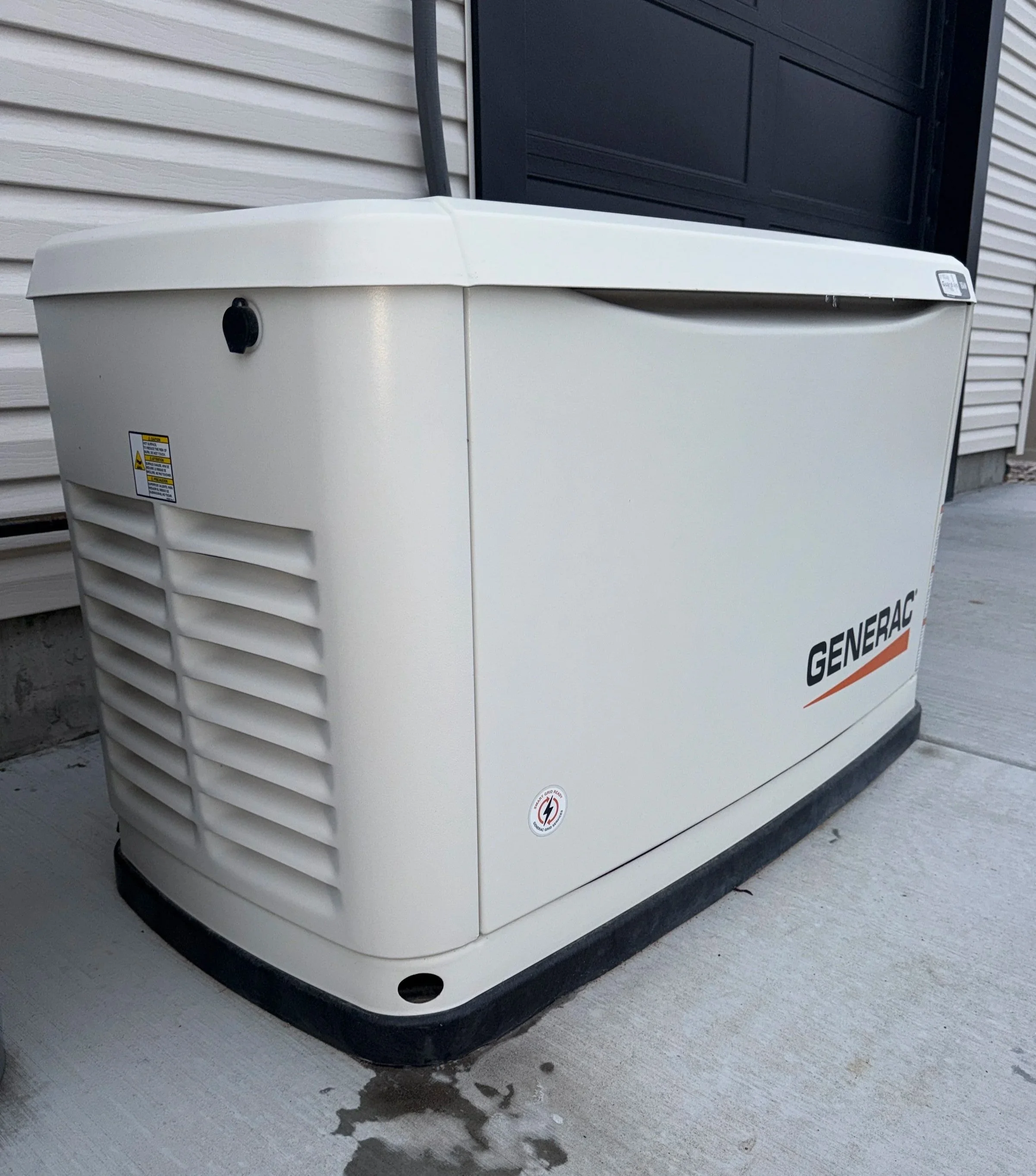 An outdoor Generac generator installed on a concrete pad next to a house with beige siding and a black garage door.