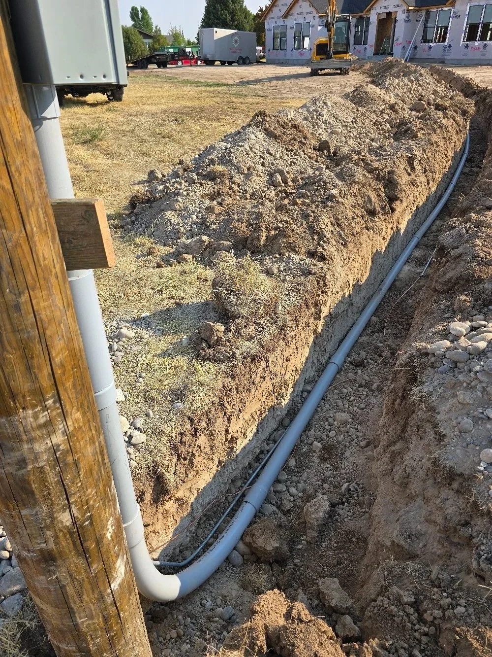 A construction site with a trench and piping, a backhoe, and an unfinished building in the background.