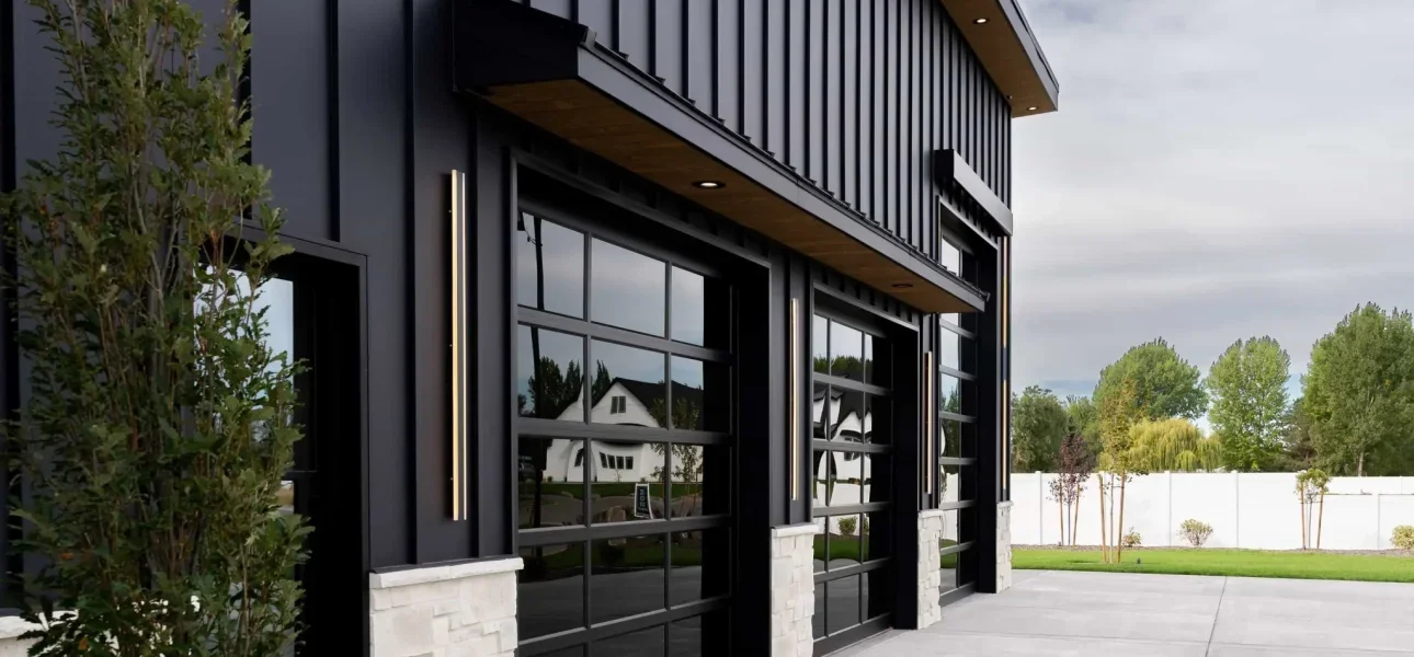 Modern building with black metal and glass garage doors, surrounded by grass and trees, with a white fence in the background.
