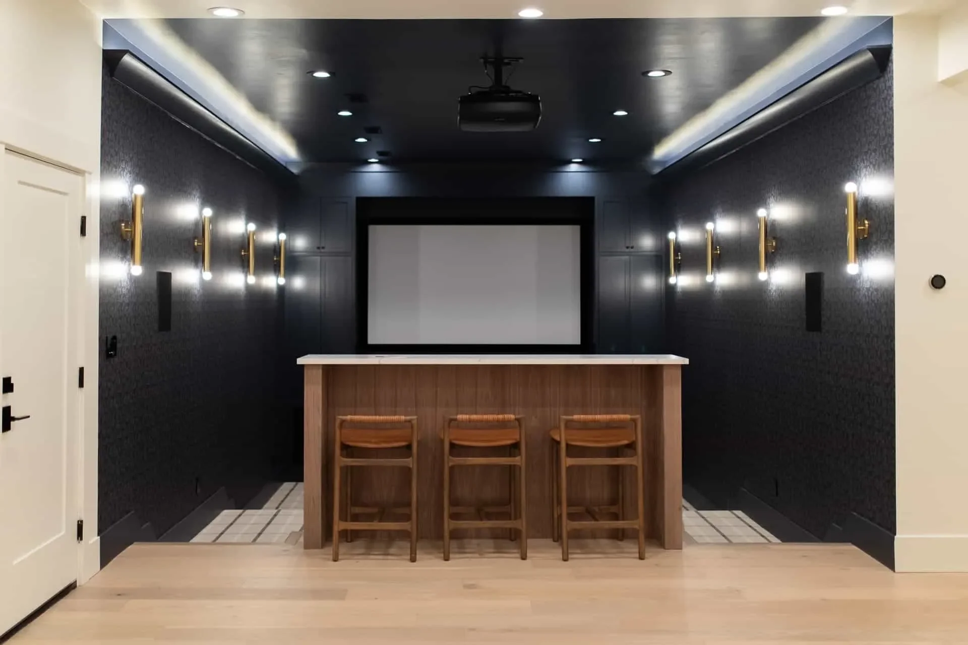 Modern home theater room with a large screen, dark walls, and wooden bar with three stools.