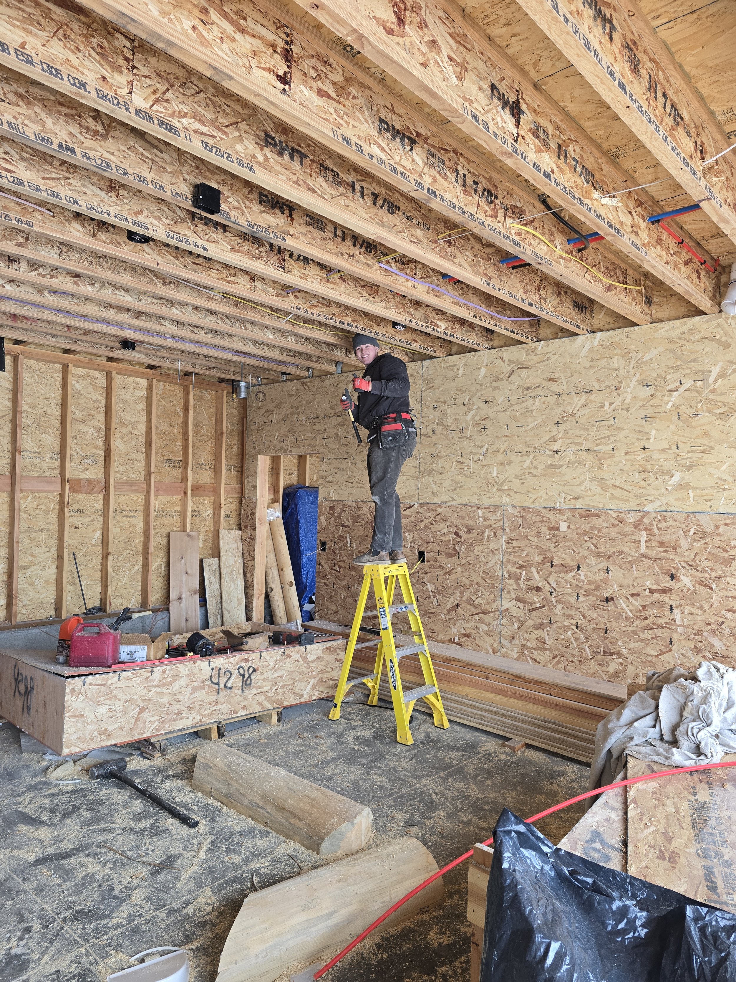 A construction worker standing on a yellow ladder working inside a building under construction, with unfinished walls and ceiling framework, tools, and building materials around.