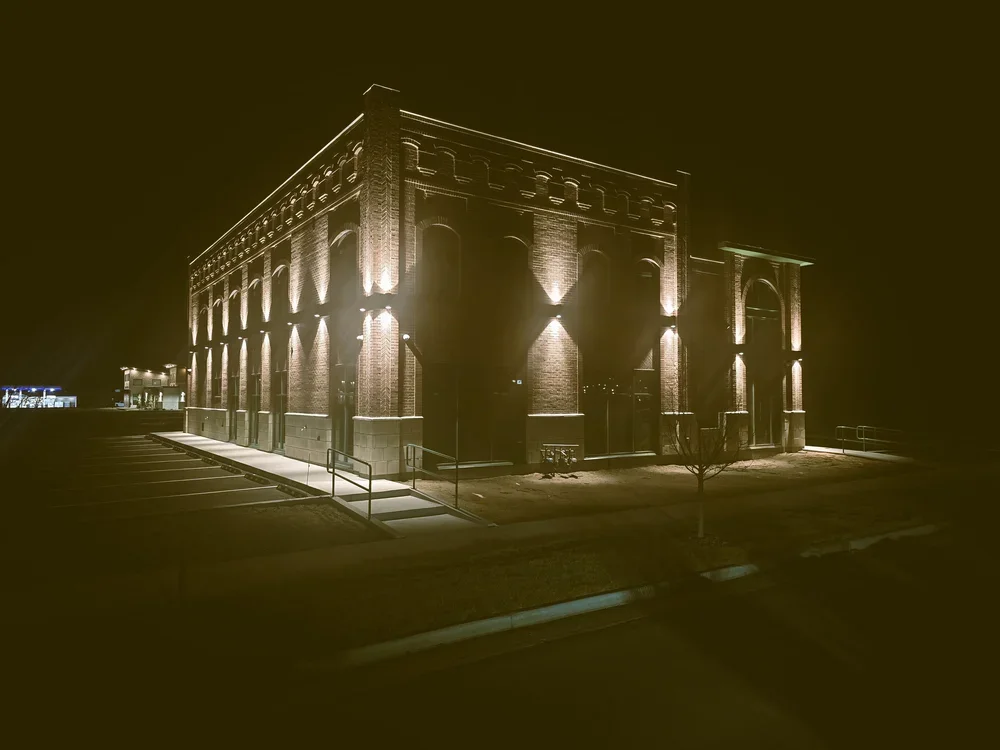 Night view of a historic brick building illuminated by exterior lights, with a parking lot and small trees in the foreground.