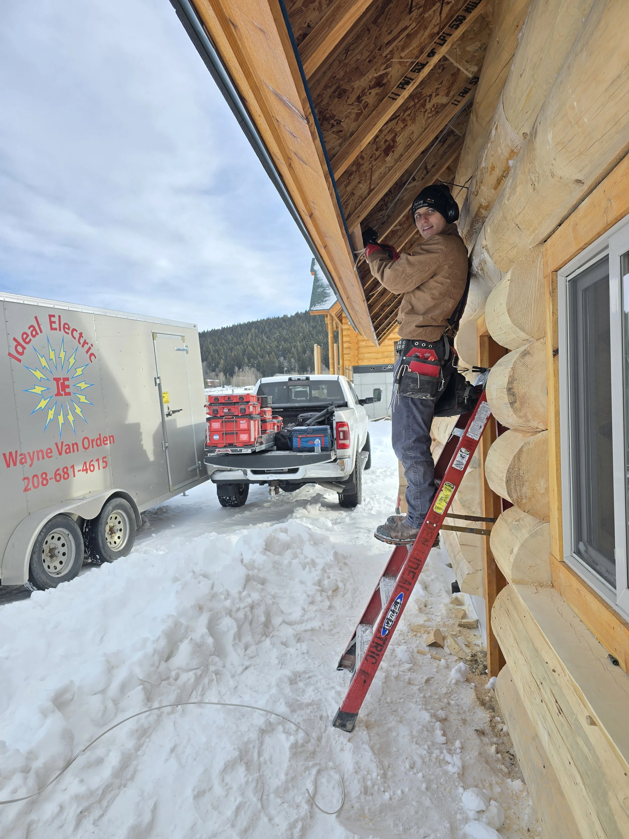 A man on a ladder installing or repairing the exterior eaves of a wooden house in a snowy landscape. There are trucks and equipment parked nearby.