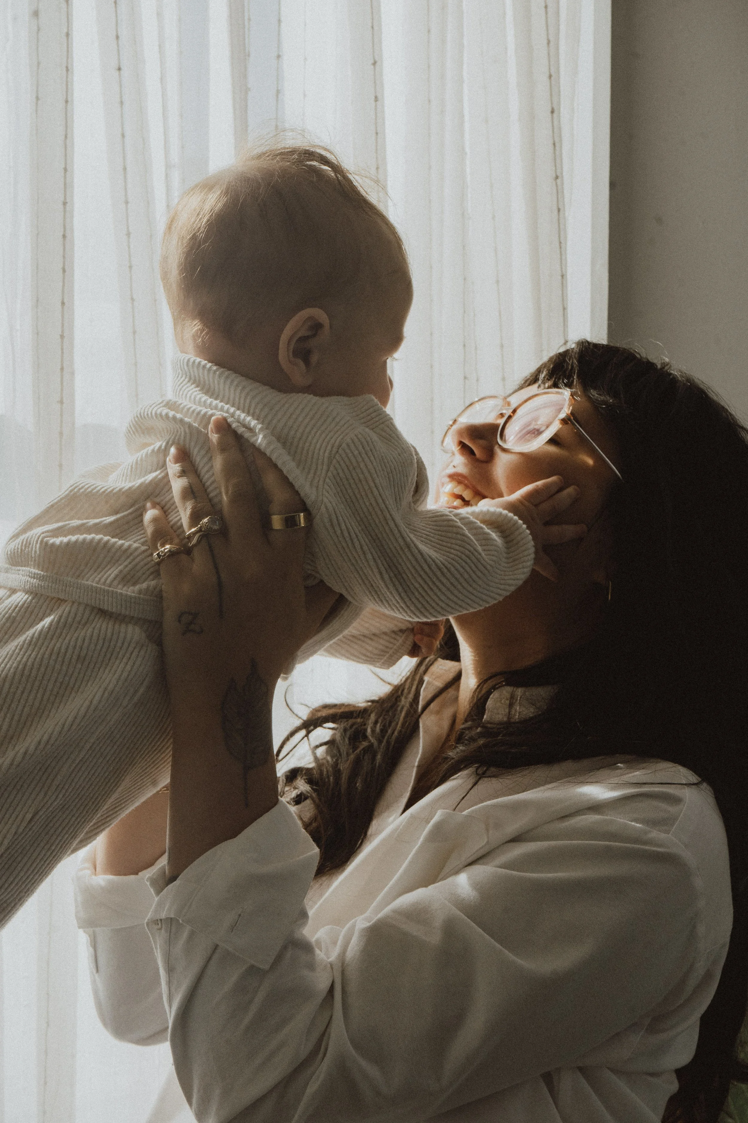 A woman with glasses holding a young child up near a window with sheer curtains, smiling and engaging with the child.