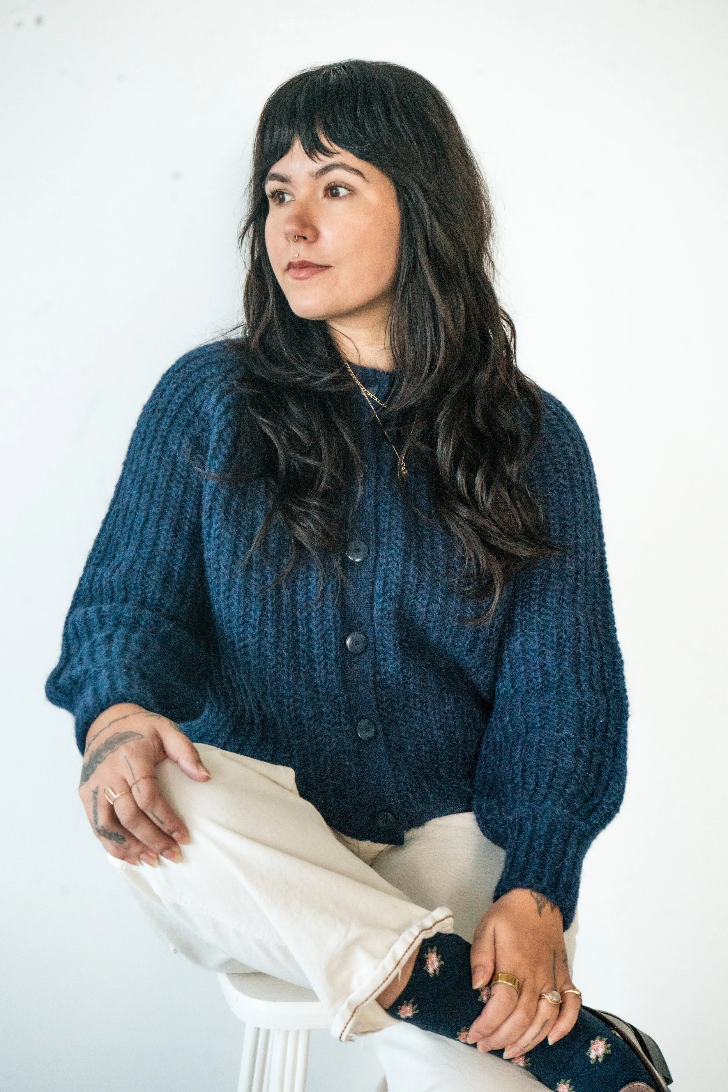 A woman with long dark wavy hair and a septum piercing, wearing a navy blue knitted cardigan, beige pants, and floral socks, sitting on a white stool against a white wall.