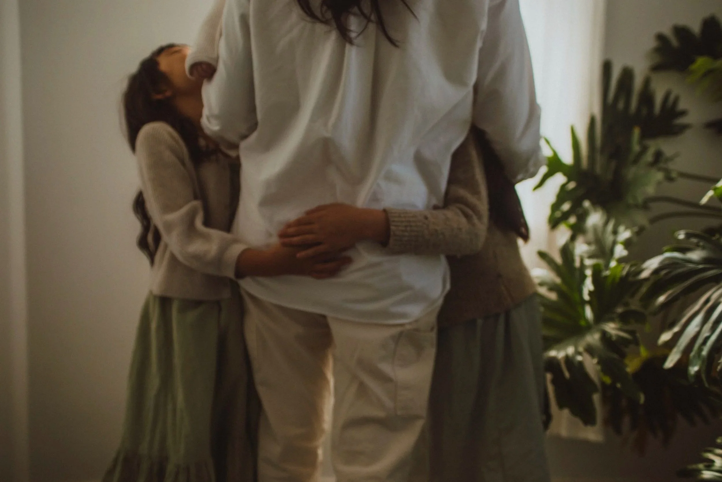 A woman in a white shirt holding a baby while hugging her two younger daughters, one on each side, in a cozy indoor setting with plants in the background.